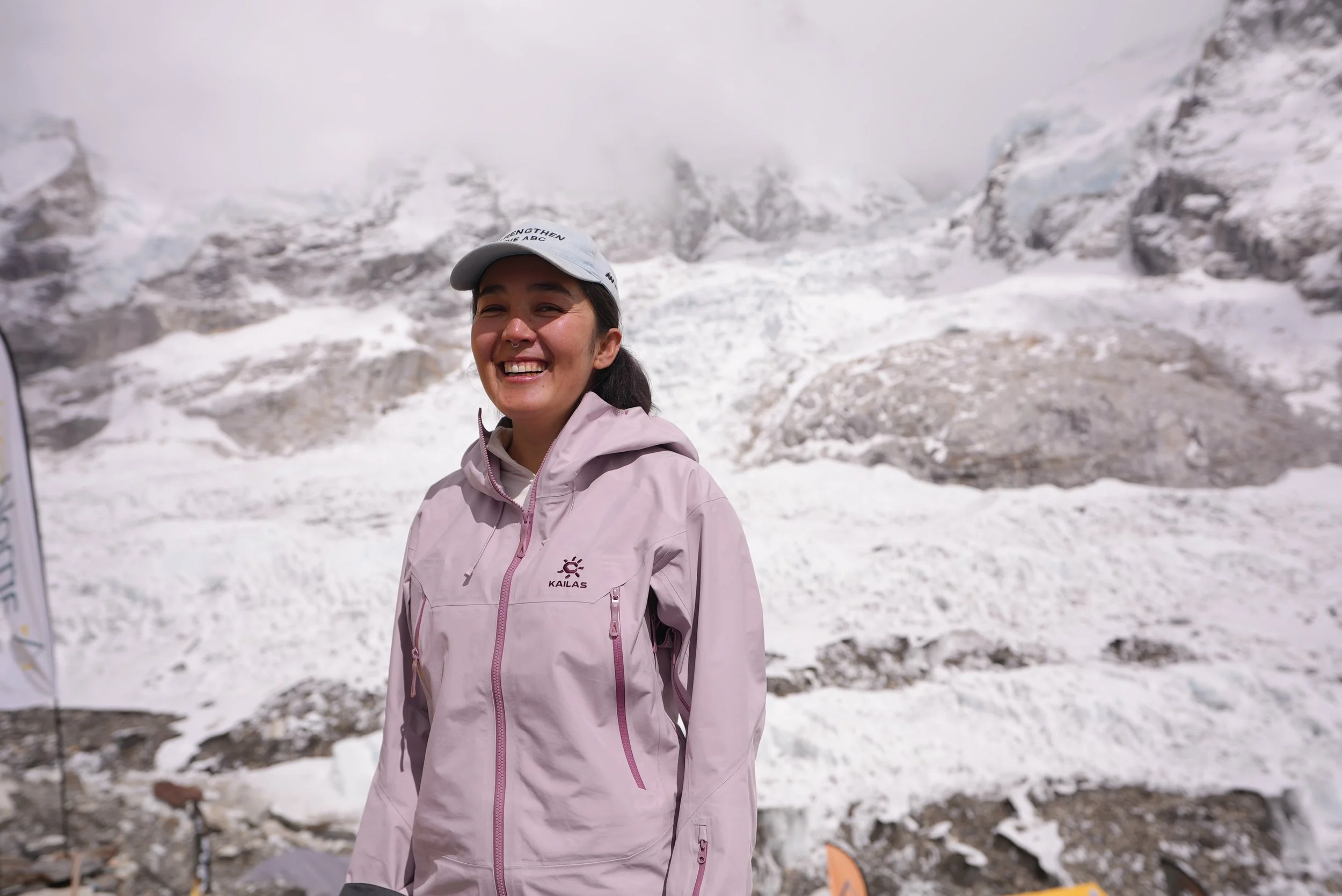 A woman smiling in a pink jacket and white cap standing on snowy mountain landscape.
