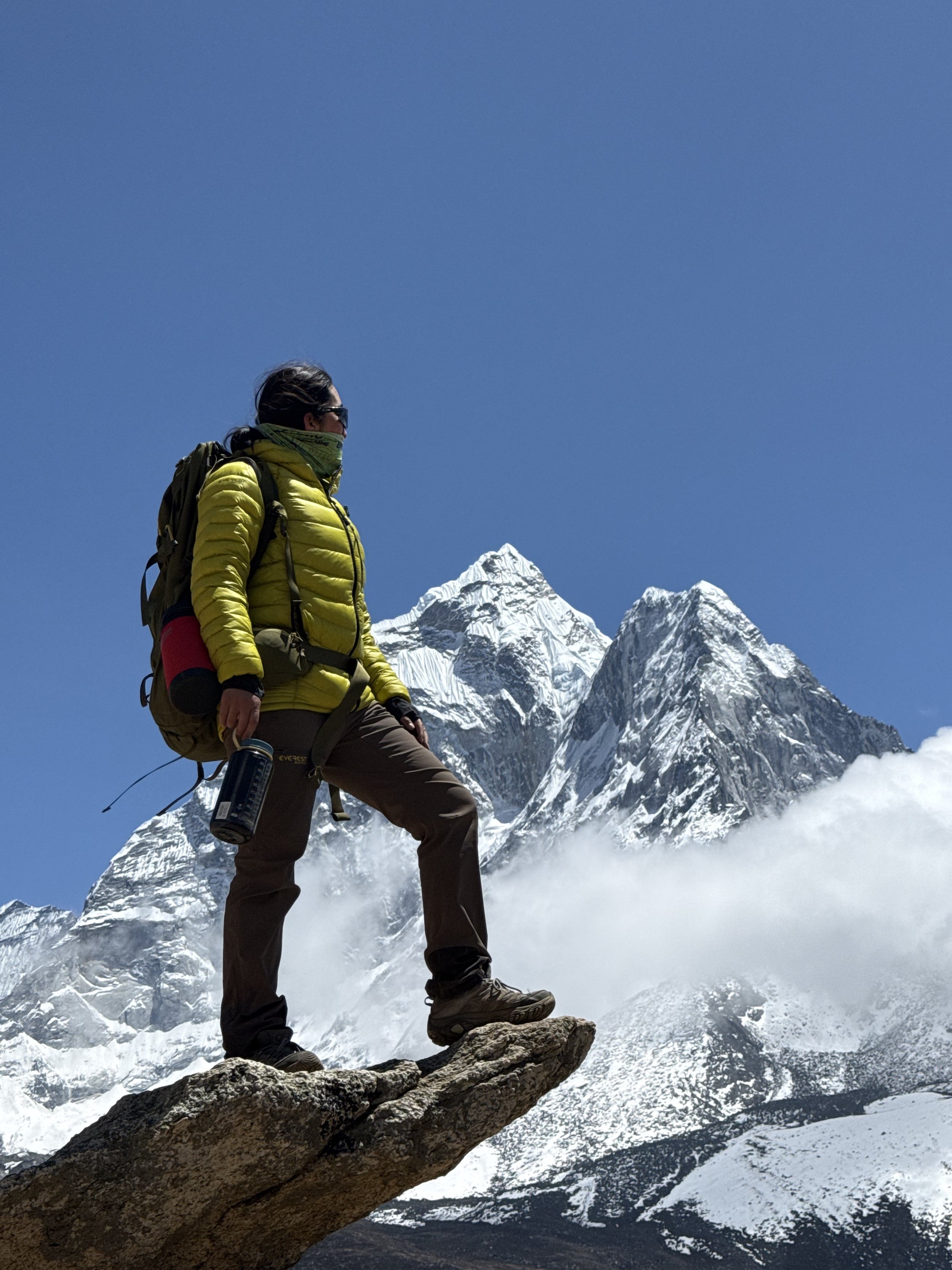 River Zakia Ahmad wearing a yellow jacket, black pants, and hiking gear standing on a rock ledge in a snowy mountain landscape with snow-covered peaks in the background.