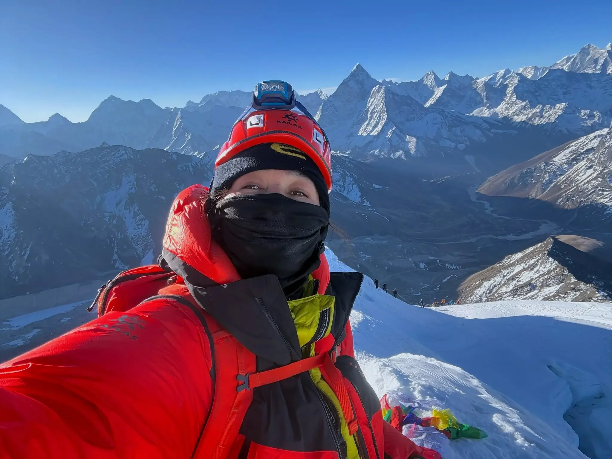River Zakia Ahmad dressed in winter snow gear, including a red jacket and black face mask, taking a selfie in a mountainous snowy landscape with snow-covered peaks and a clear blue sky in the background.