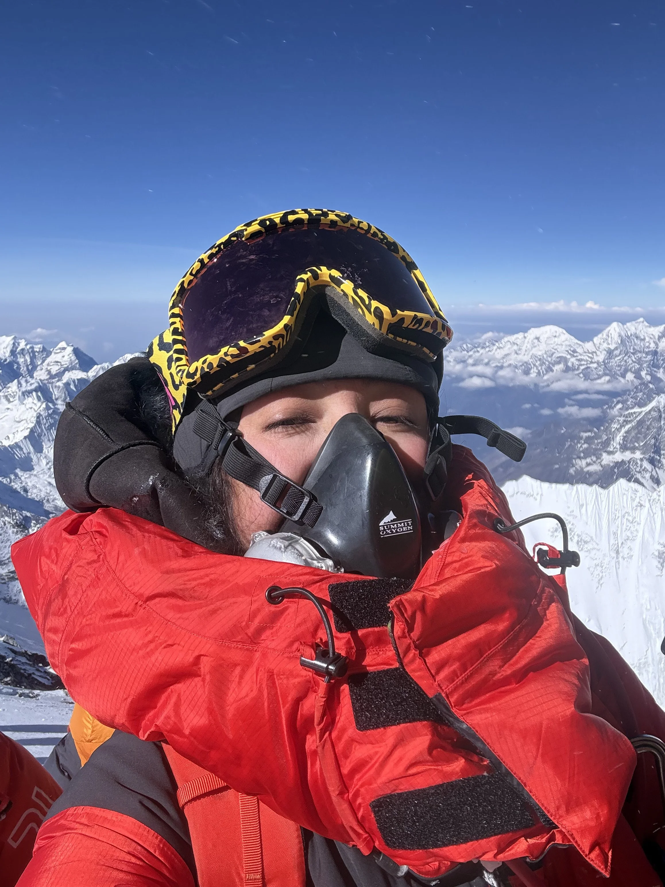River Zakia Ahmad taking a selfie in snowy mountain terrain, wearing a red jacket, a black helmet, black gloves, and yellow sunglasses, with snow-capped mountains in the background.