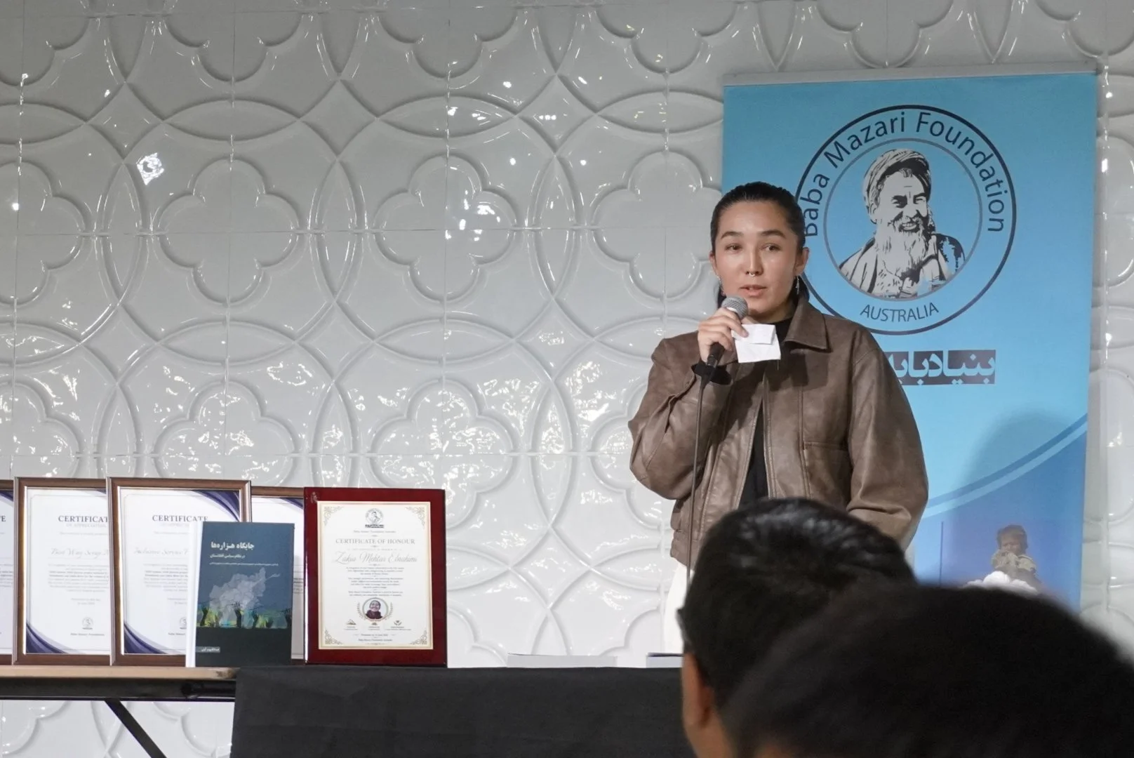 A young woman with dark hair tied back, wearing a brown leather jacket, is holding a microphone and speaking at an event. Behind her is a blue banner with a logo for the Baba Mazari Foundation Australia, featuring a cartoon illustration of an older m