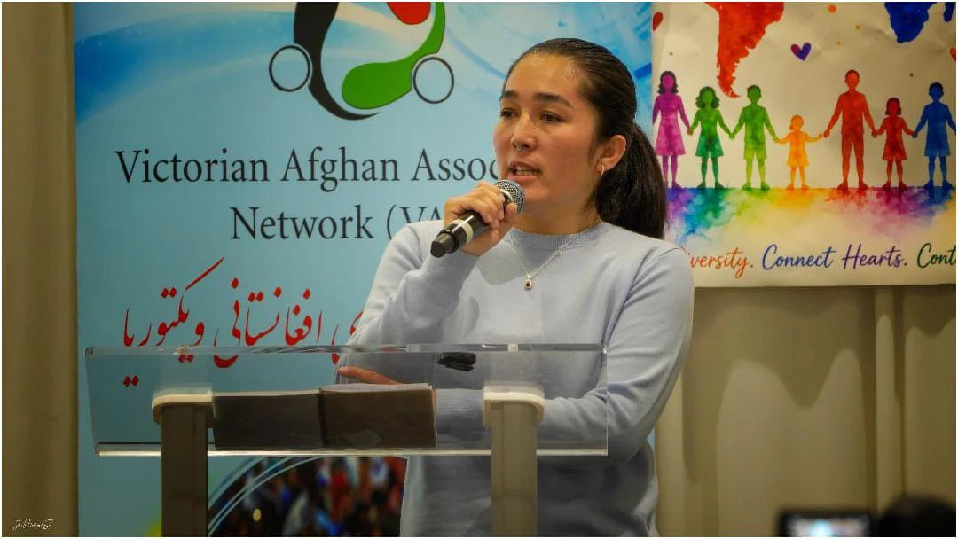 A woman speaking at a podium during an event for the Victorian Afghan Association Network, with a colorful banner in the background showing a rainbow of people holding hands and the words 'Diversity. Connect Hearts. Continue'.