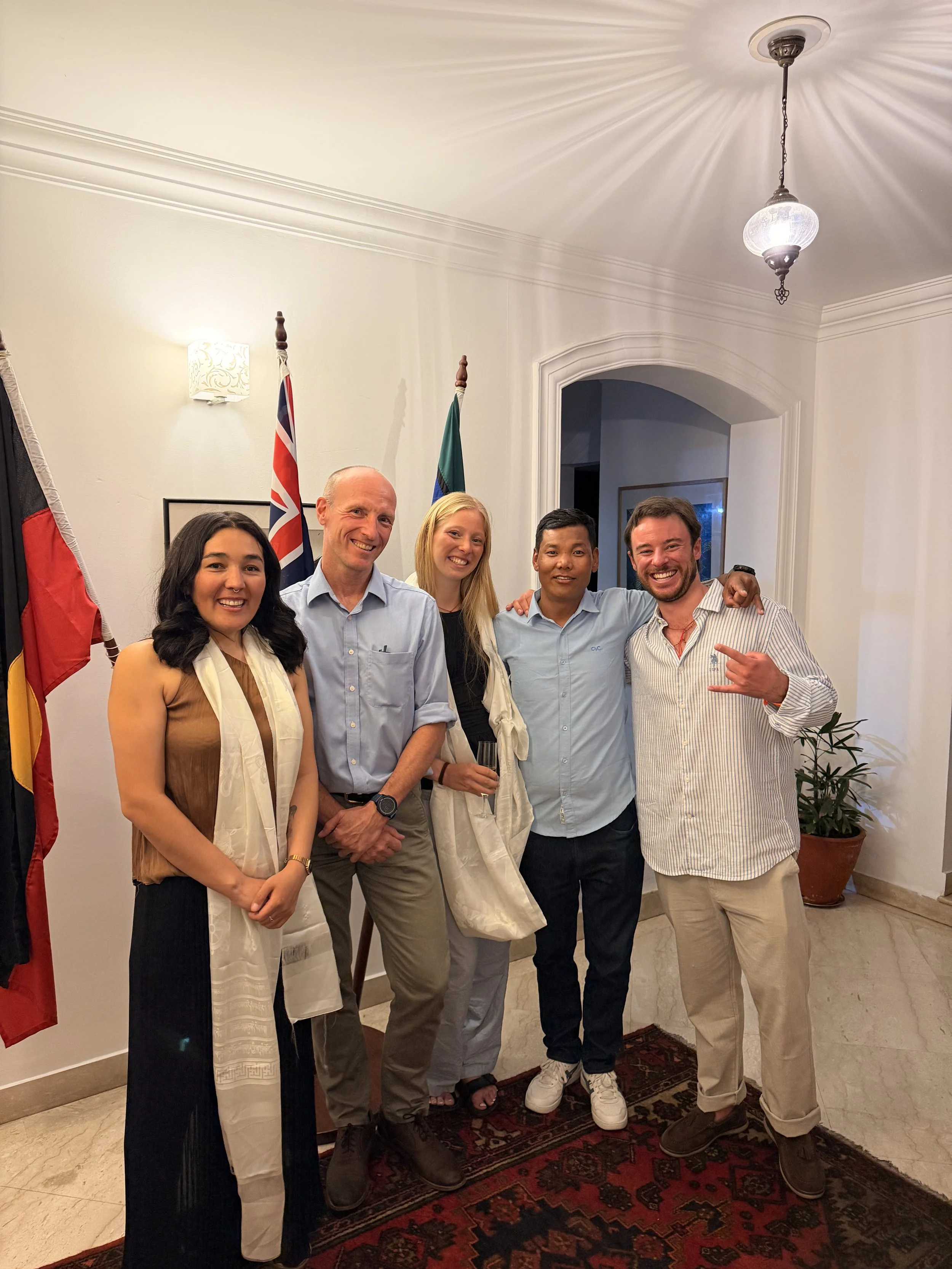 Group of five people smiling and standing close together indoors with flags and decorative lighting in the background.