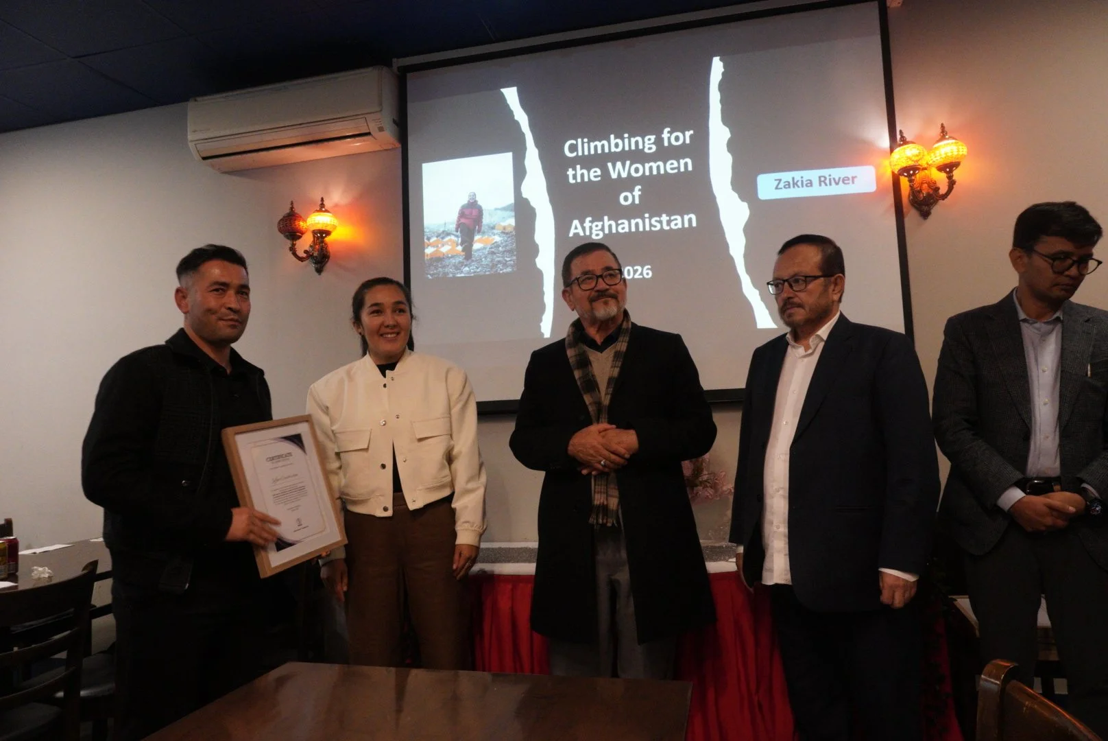 A group of five people standing in front of a presentation screen. The screen displays the title 'Climbing for the Women of Afghanistan' along with an image of a woman climbing. One person is holding a framed certificate. The room has wall-mounted la