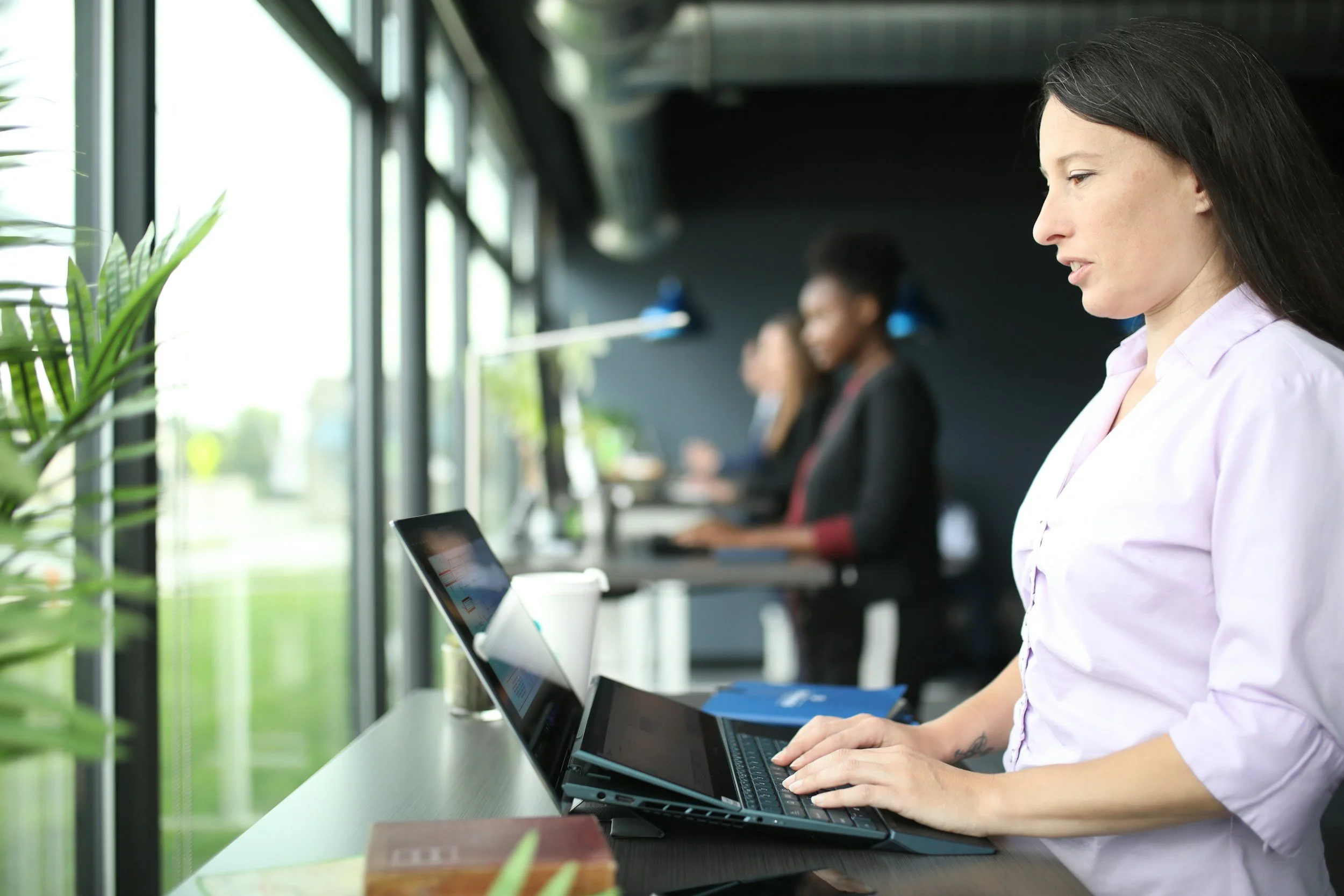 Woman using a laptop in a modern office with large windows and green plants, with two other women in the background also working.