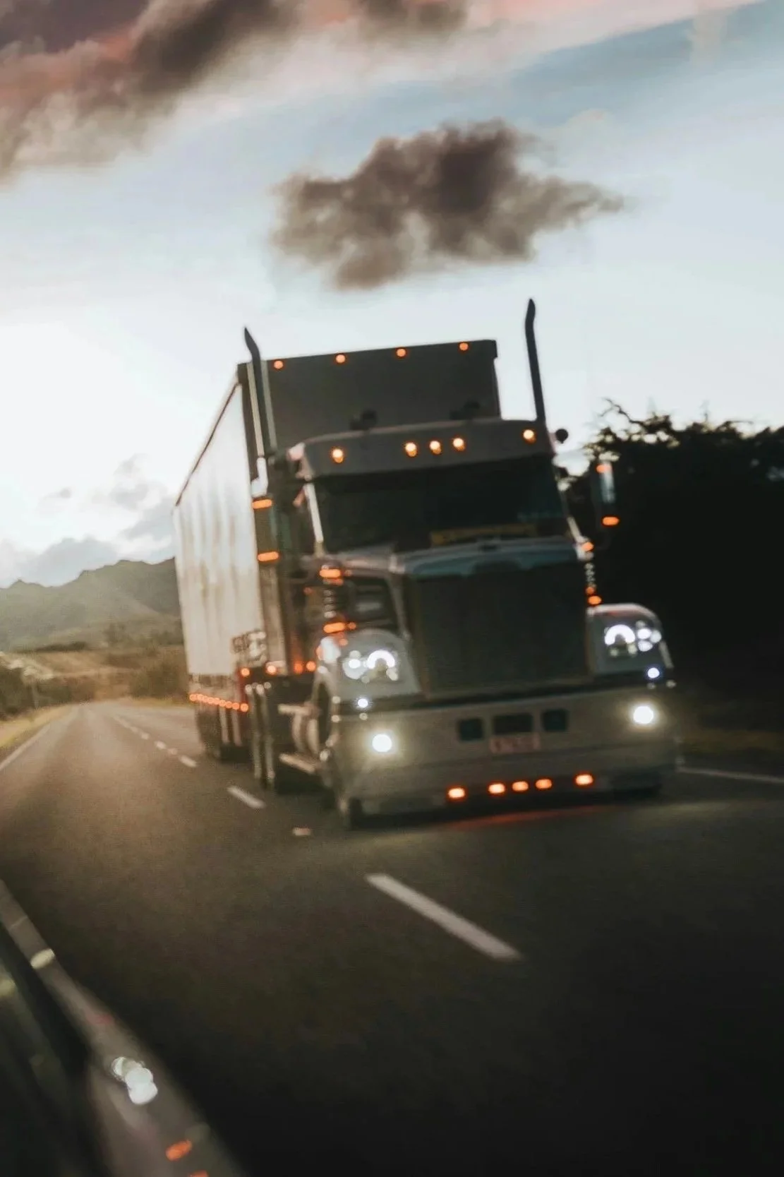 A large semi-truck with a dark blue cab and white trailer driving on a highway during dusk with mountains in the background and dark smoke rising into the sky.