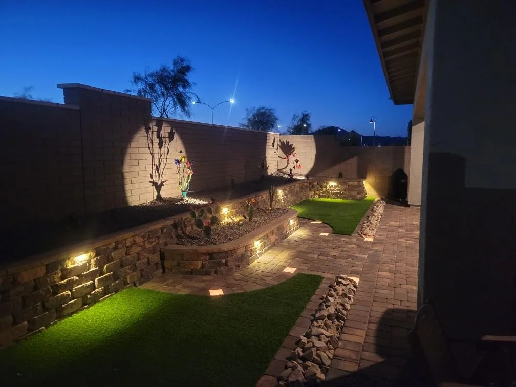 Backyard patio at night with brick walkway, lit flowerbed with metal sculptures, cacti, and decorative landscape lighting.