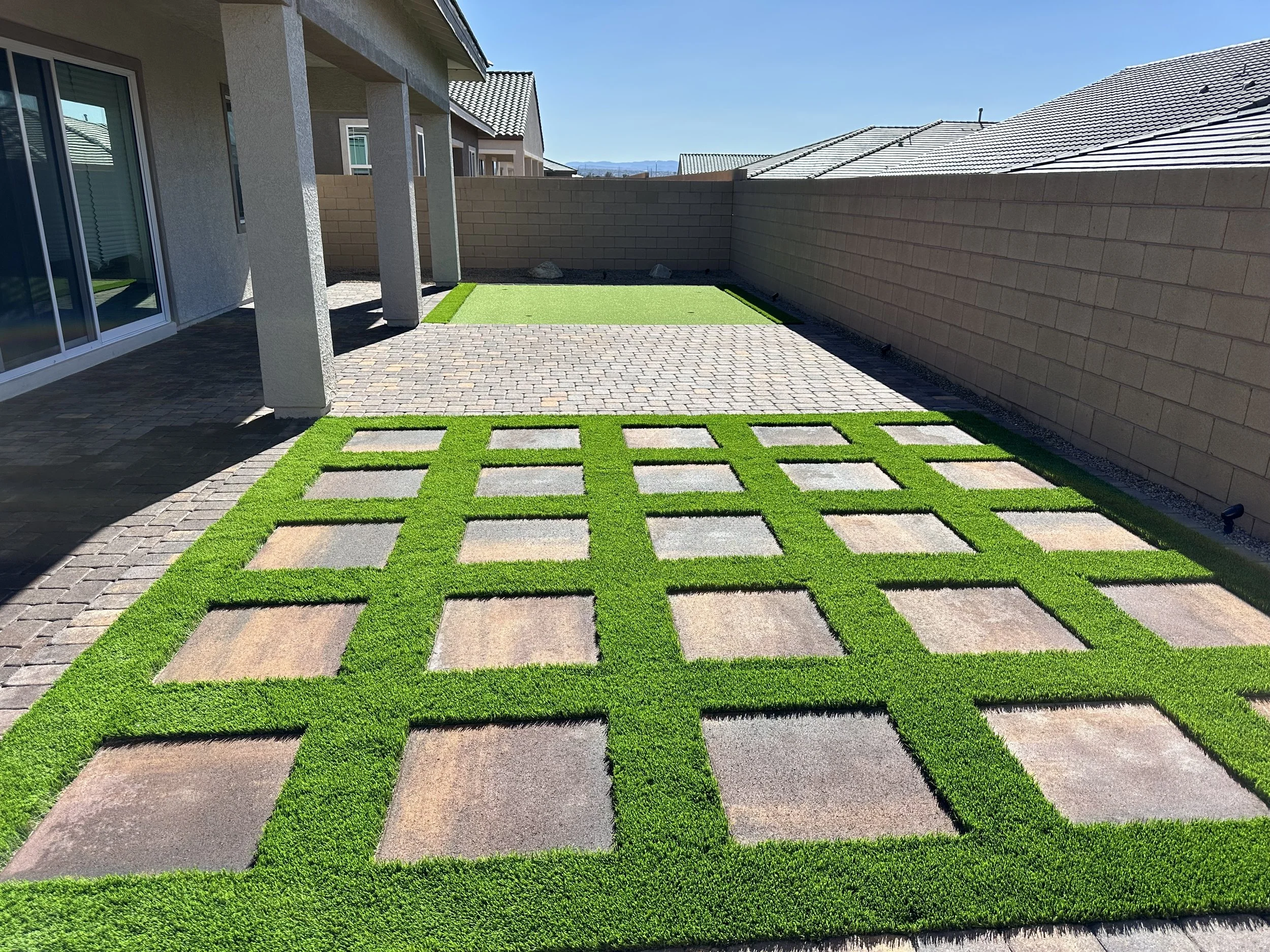 Patio area with a paved brick surface and artificial grass squares, enclosed by beige brick walls, under a clear blue sky.