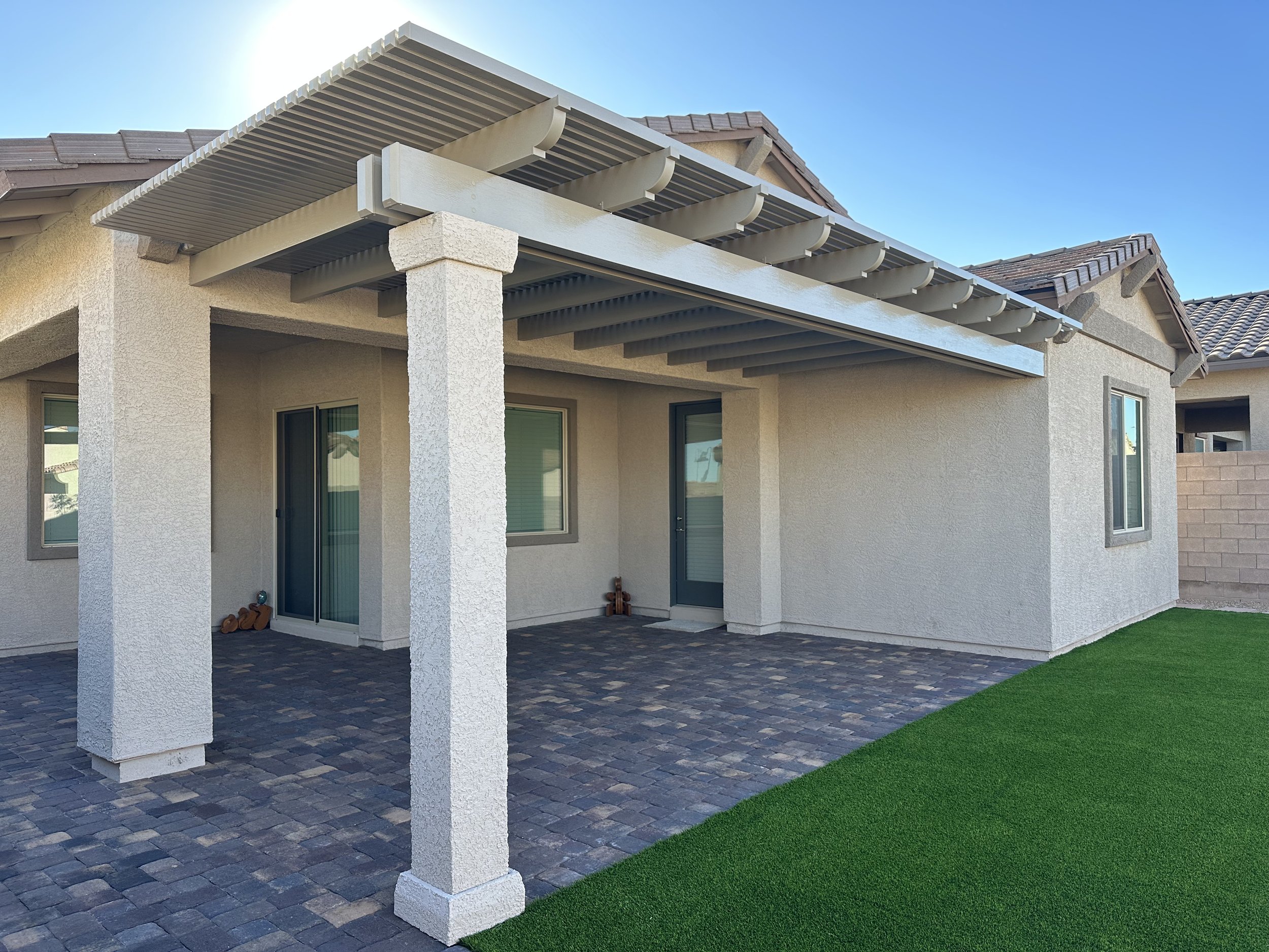 Back patio of a house with a covered pergola supported by textured white columns, a tile roof, sliding glass doors, a brick wall, a paved patio area, and a strip of artificial grass.