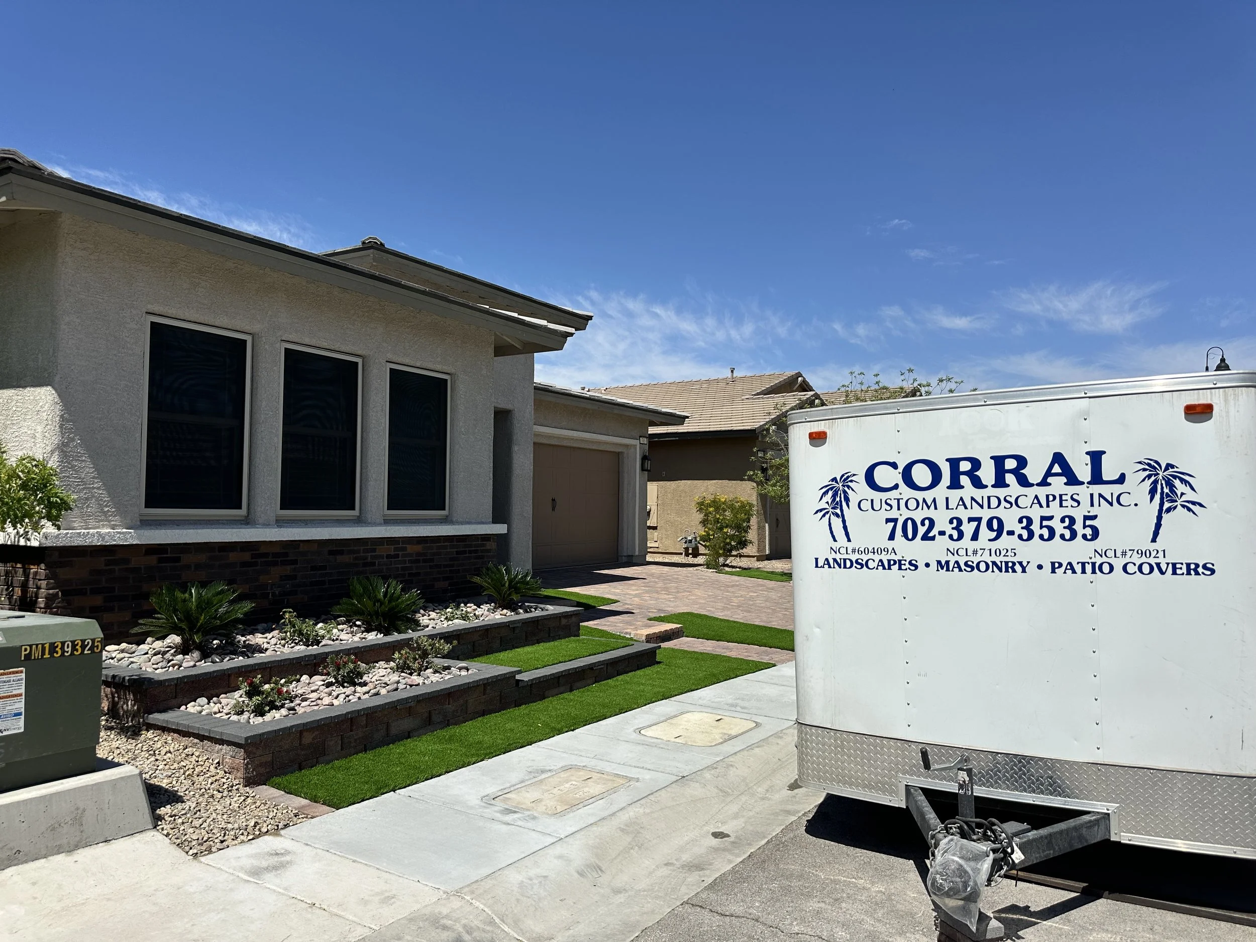 A residential front yard with a landscaped garden bed filled with white rocks, small green plants, and a stone border. There are three windows on the house's exterior wall, a driveway, and a small tree. A large white trailer with blue and black lettering in the front yard belongs to a landscaping company called Corral Custom Landscapes Inc.