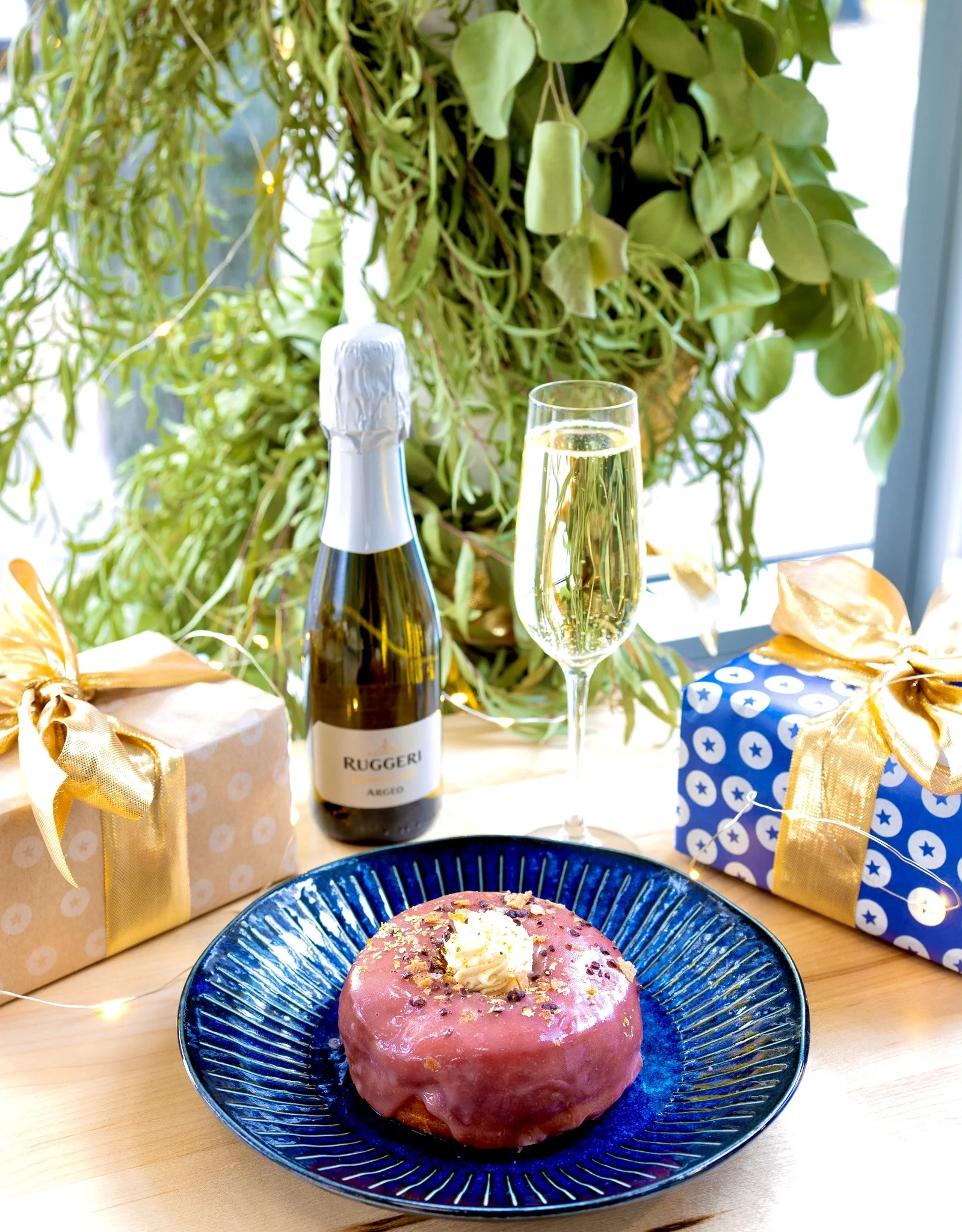 A pink frosted donut with sprinkles and white icing on a blue plate in the foreground. Behind it, there is a bottle of sparkling wine, a glass of champagne, and two wrapped gift boxes with gold and blue wrapping paper and gold ribbons. In the background, there are green plants and fairy lights.