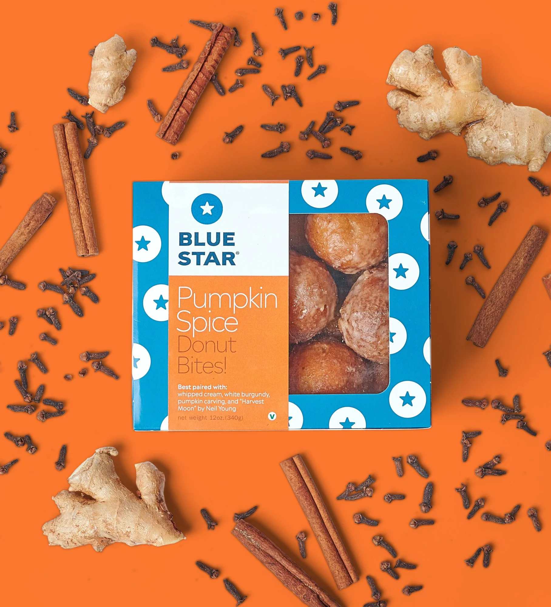 Top view of a box of Blue Star Pumpkin Spice Donut Bites with ginger, cinnamon, and cloves scattered around on an orange surface.