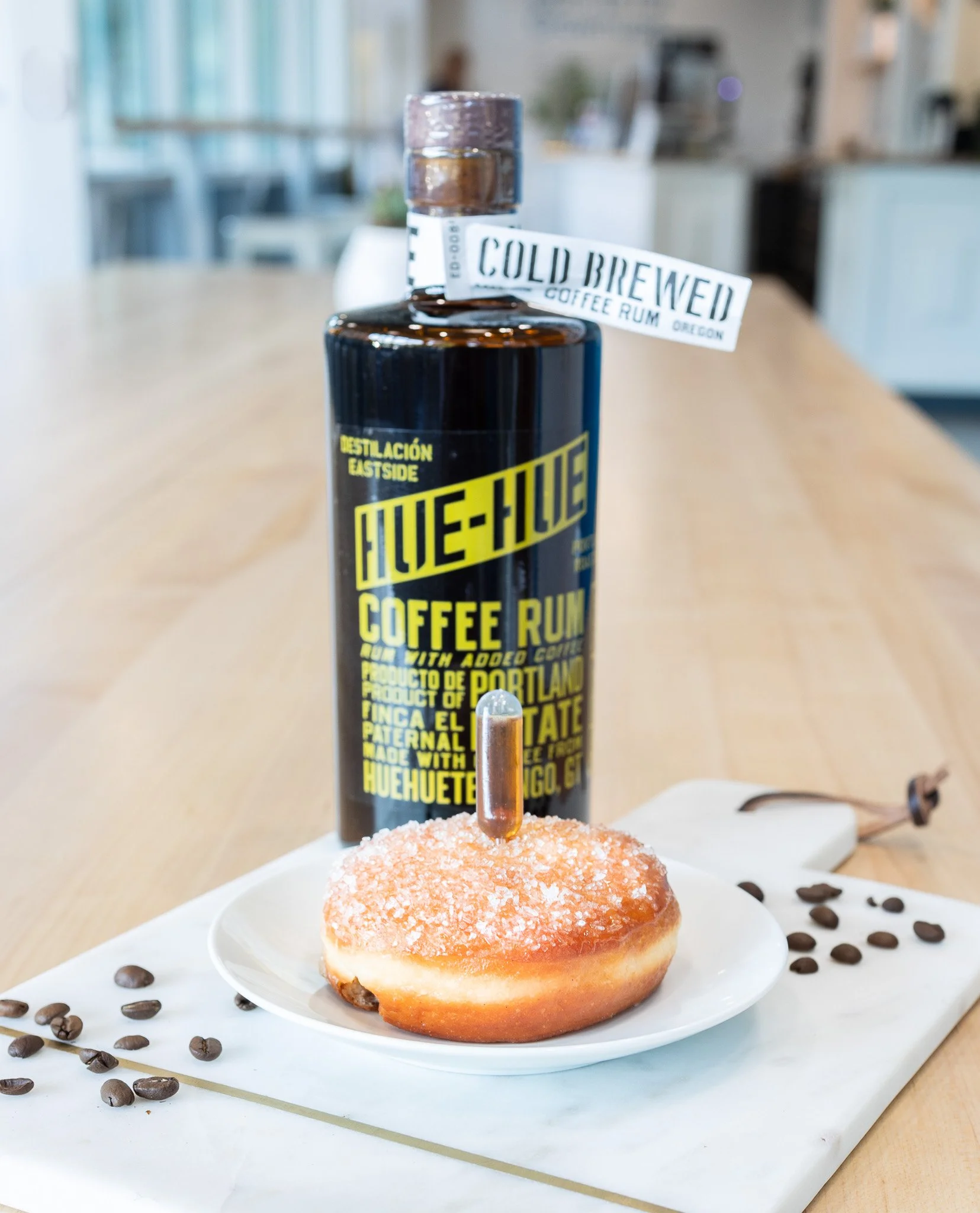 Close-up of a vanilla donut with sugar crystals on top, with a hot shot coffee bottle with a tag that reads 'Cold Brewed Coffee Rum' placed behind it. The donut is on a white plate, decorated with scattered coffee beans on a light-colored wooden table.