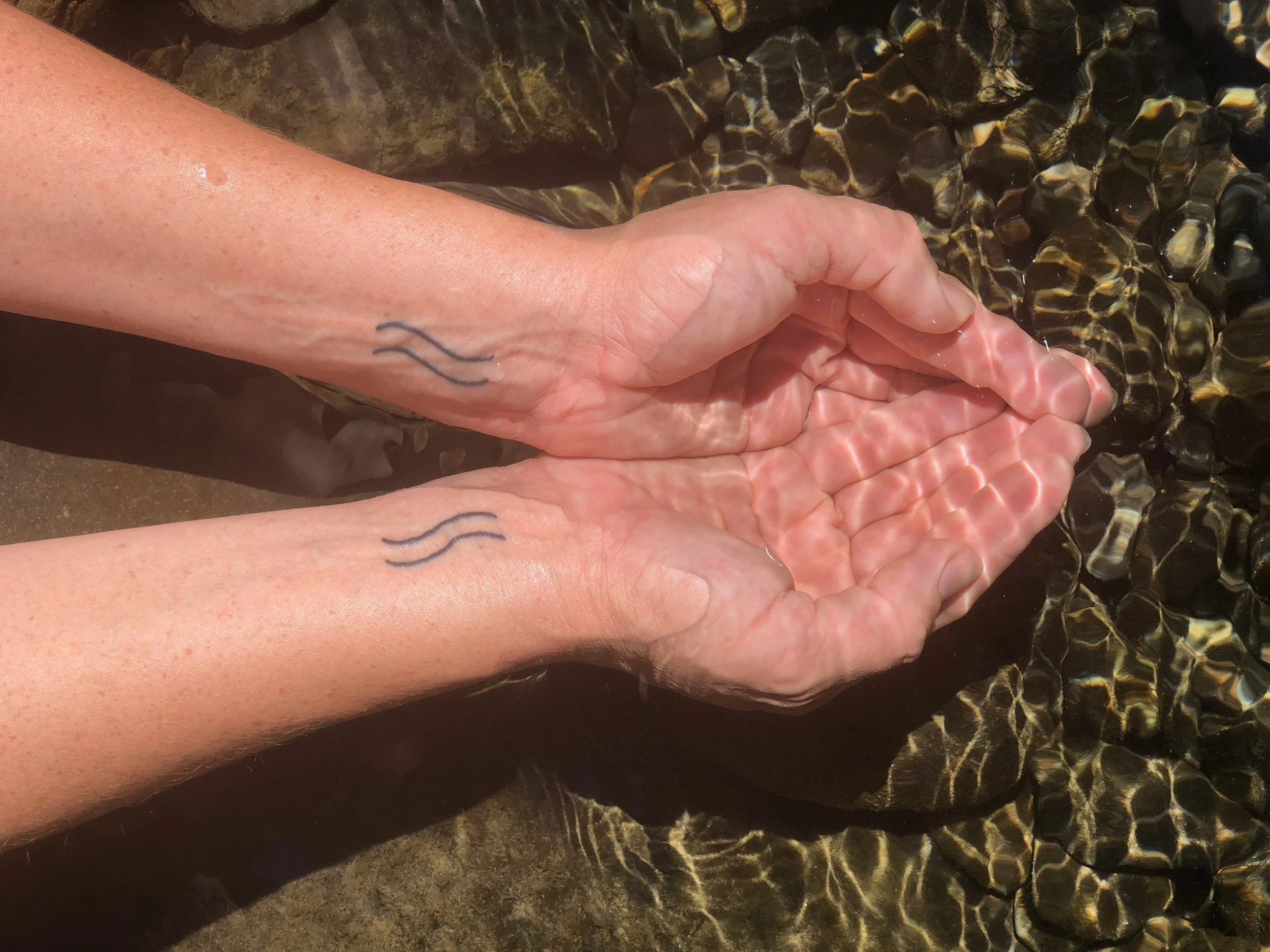 Person with light skin and two tattoos on the wrist, reaching out with hands submerged in shallow clear water over rocks at the beach.