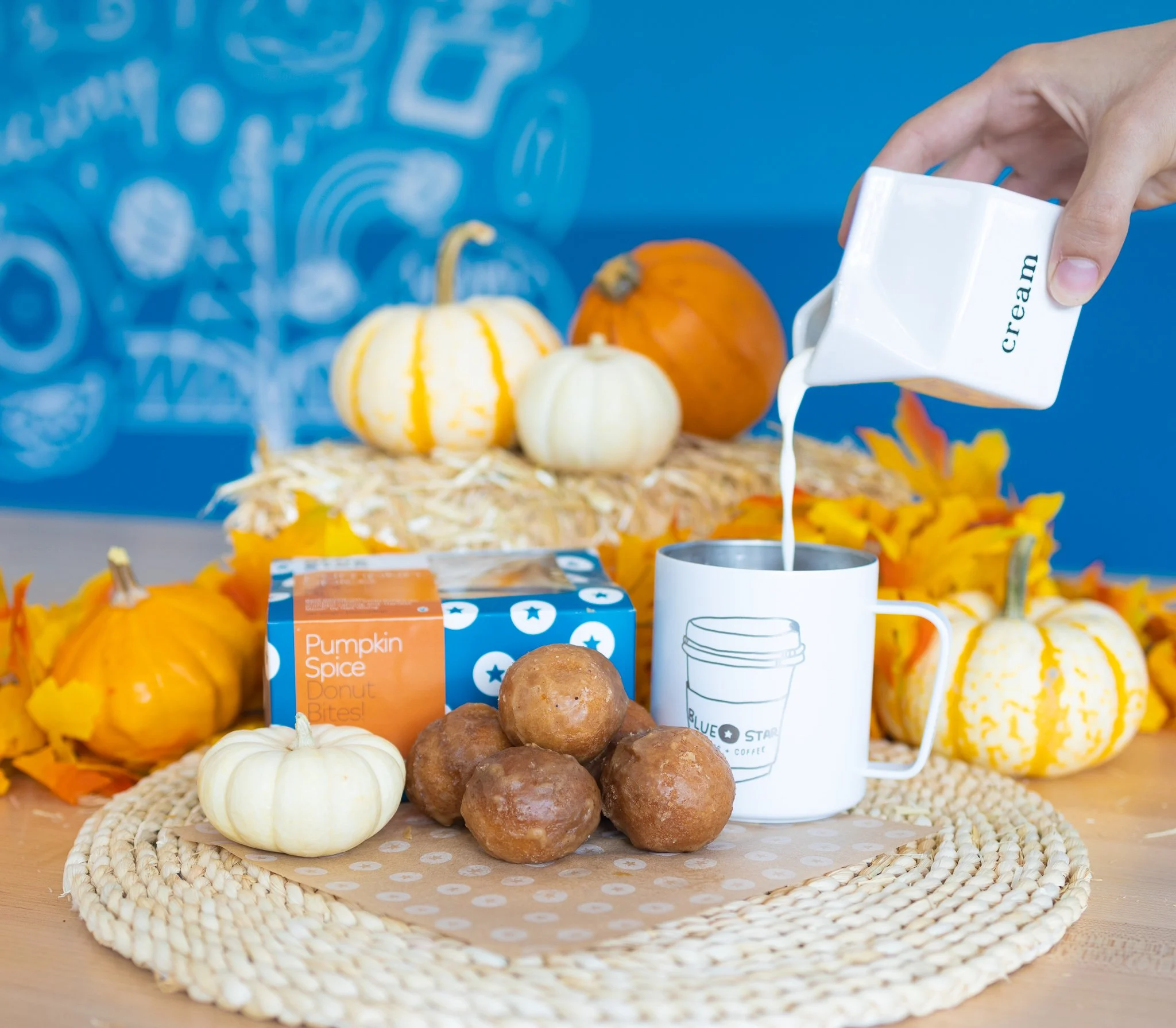 Various pumpkins and a paper cup with a coffee illustration, arranged on a woven mat and a wooden surface with a blue background.