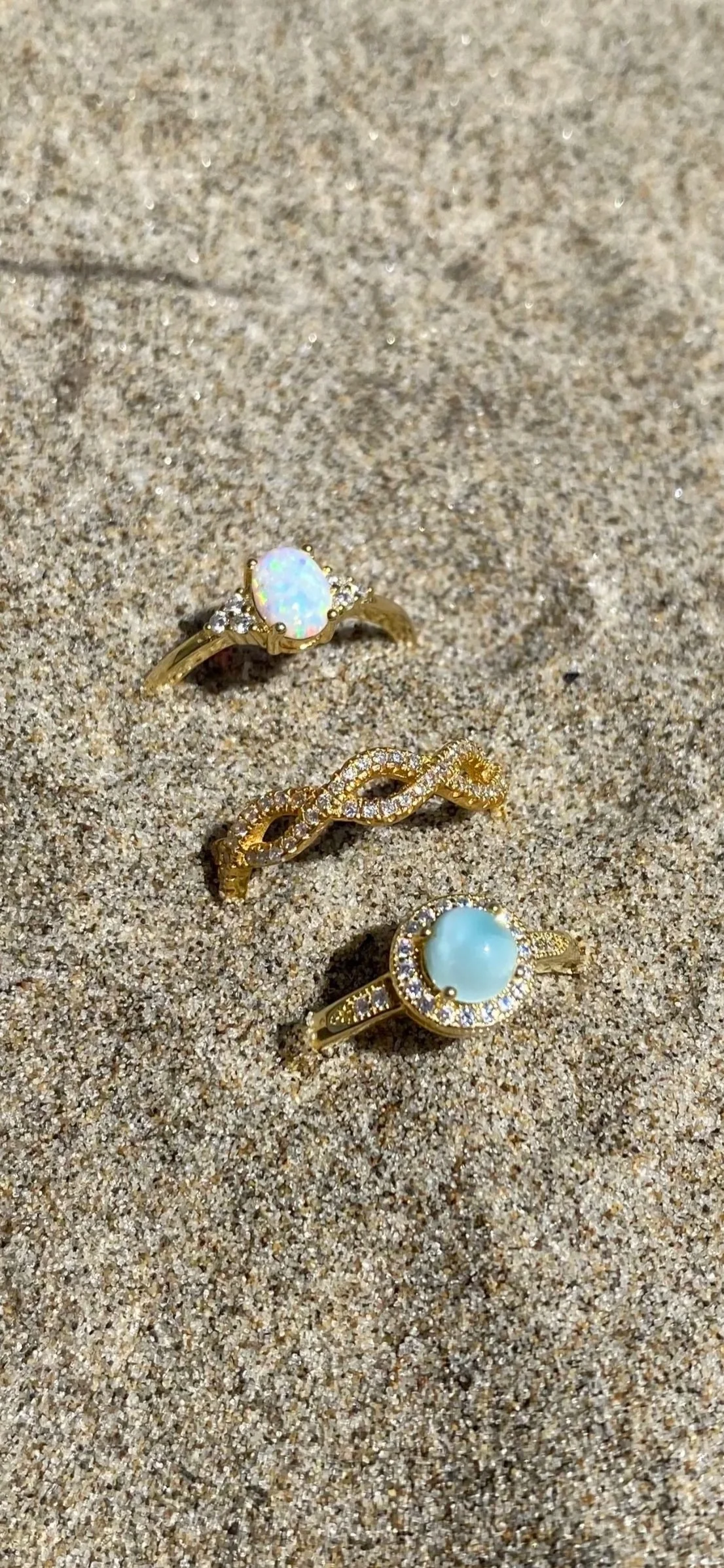 Three gold rings with large pearls and diamonds resting on sand.