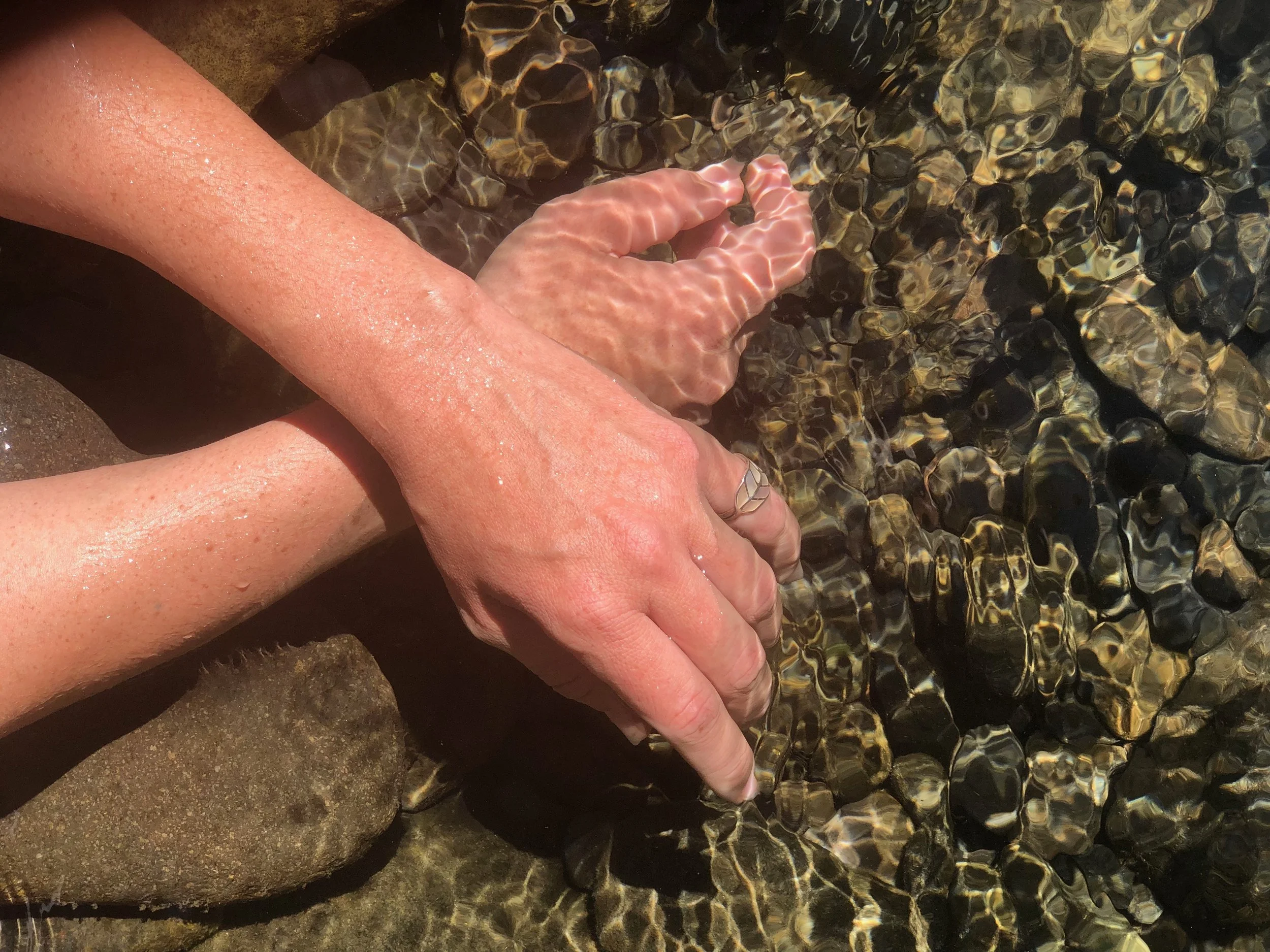 Person’s arm and hand gently touching shallow water over rocks and pebbles on a beach or creek.