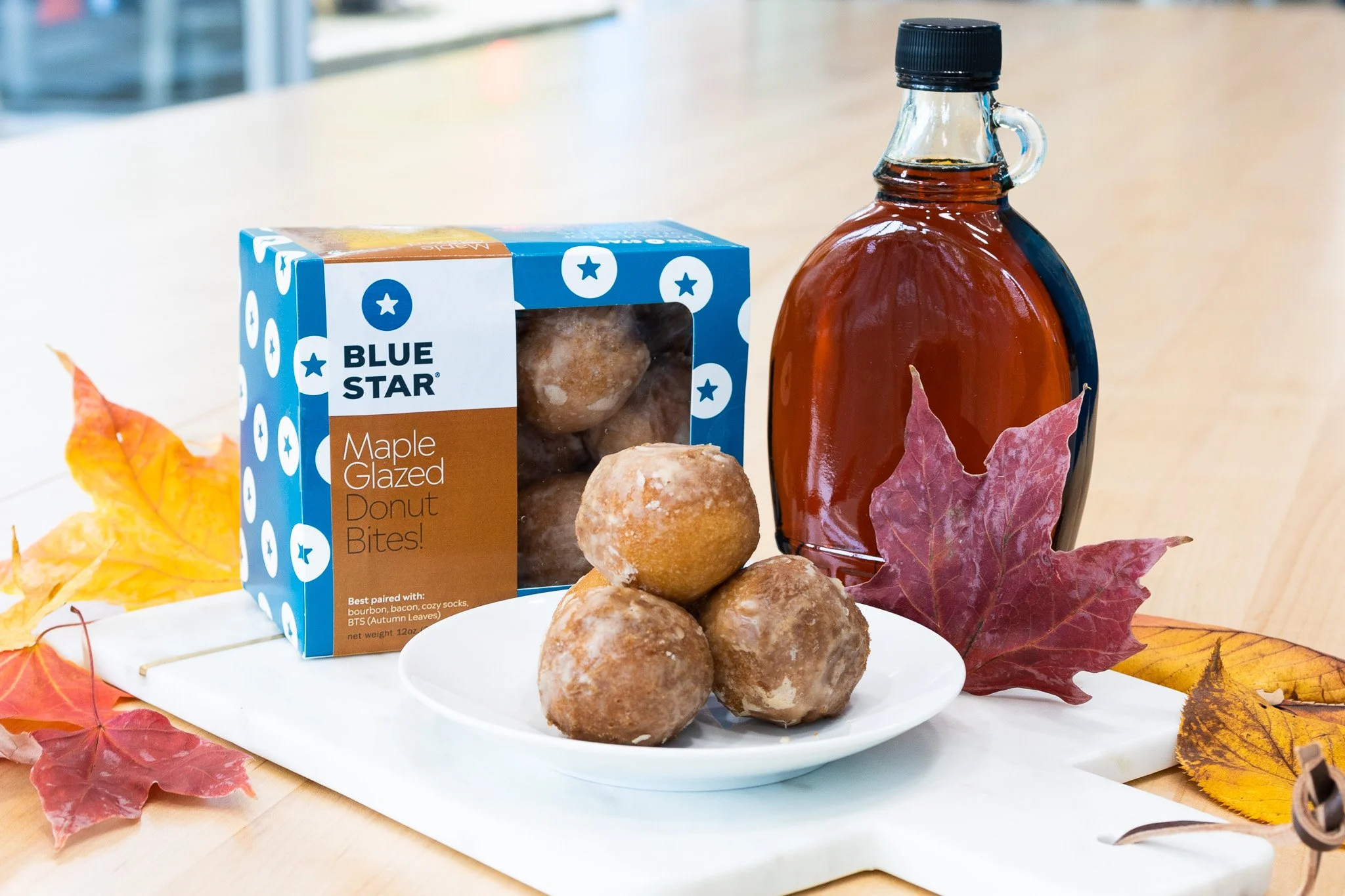 Plate of glazed donut bites next to a box labeled Blue Star Maple Glazed Donut Bites, with autumn leaves and a bottle of syrup in the background.