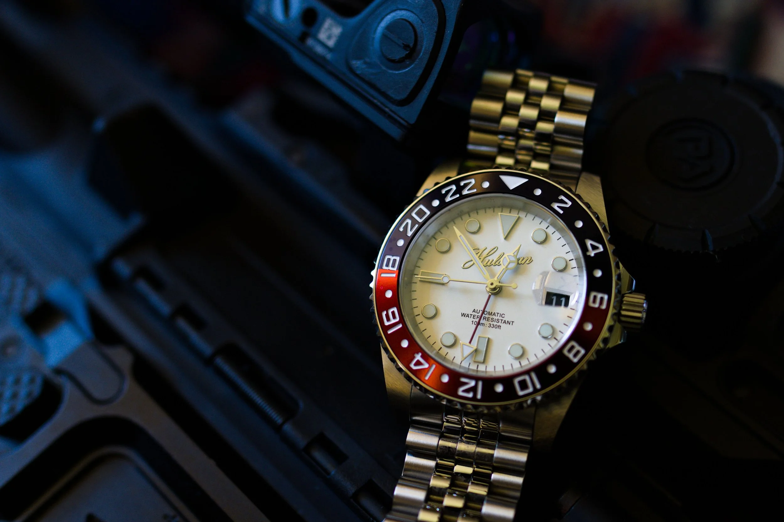 A close-up of a wristwatch with a silver band, cream-colored dial, and black-and-red bezel, resting on a surface with electronic components.