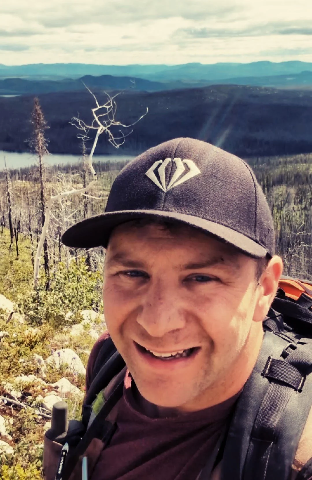 A man smiling outdoors in a mountainous area with a backpack, wearing a black cap with a diamond emblem, and surrounded by trees and scenic landscape in the background.