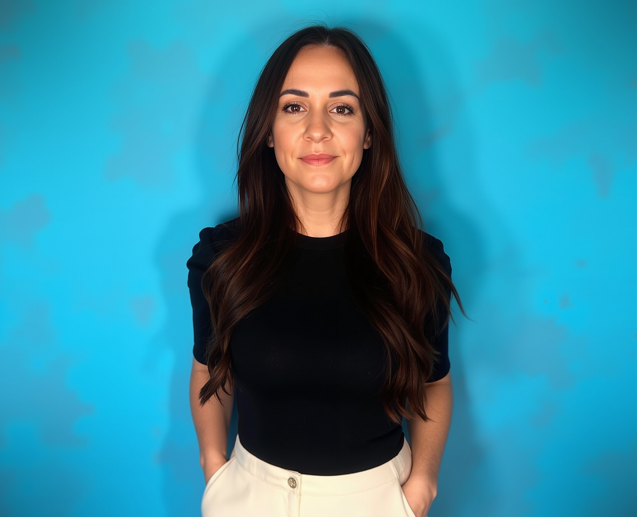 A woman with long dark hair wearing a black top and white pants standing against a solid blue background.