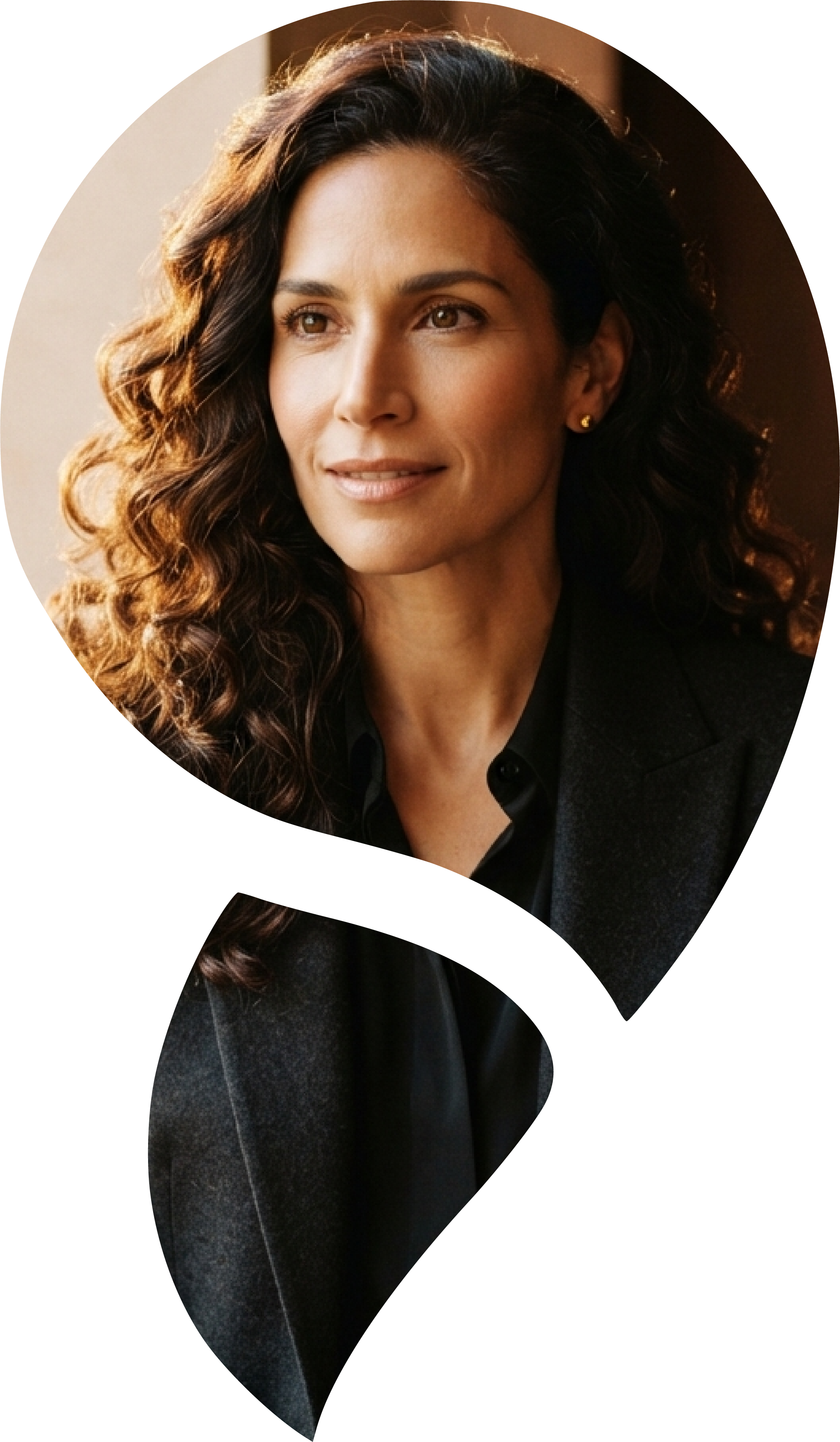 A woman with wavy brown hair wearing a dark blazer and stud earrings, sitting in front of a window with warm lighting.