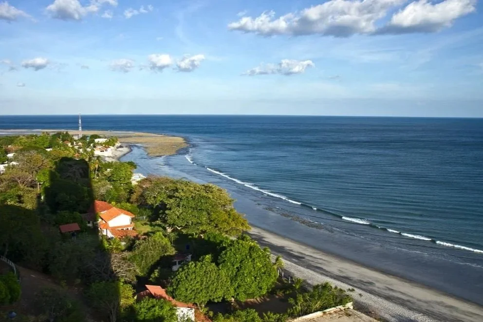 En kystlinje med sandstrand og bølger, omgitt av grønne trær og noen hus i en fjernliggende landsby. Havet er blått og himmelen er delvis overskyet.