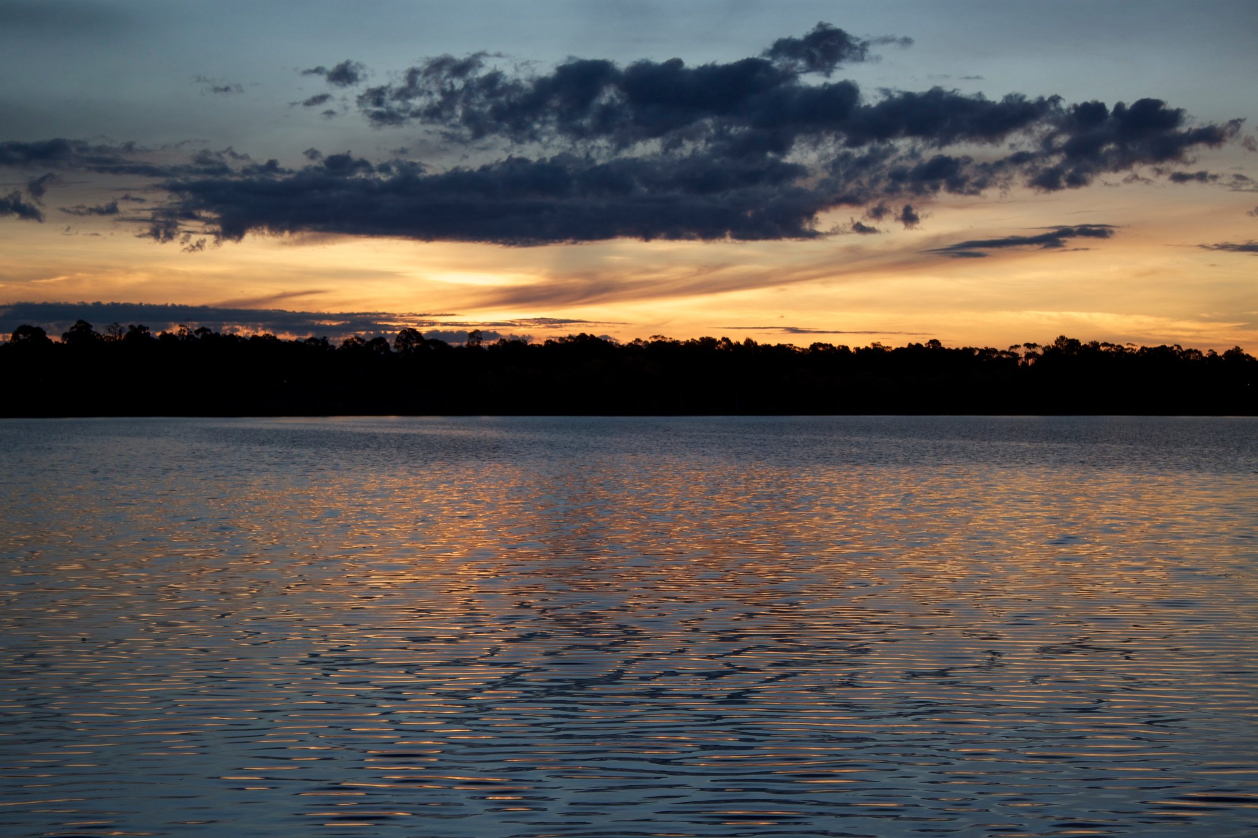 Sunset over Lake Ginninderra in Canberra with dark clouds in the sky and a silhouette of trees on the horizon.