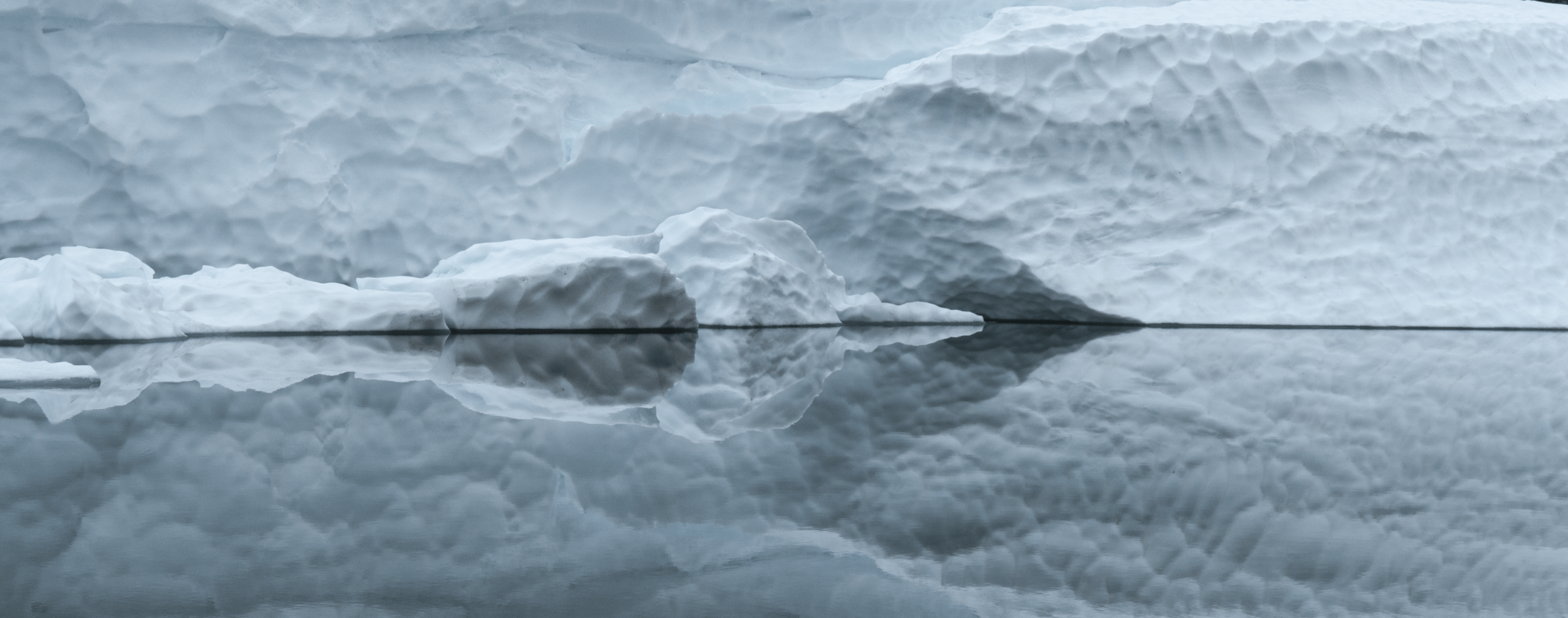 A large piece of ice floating in calm water, with its reflection visible on the water's surface, set against a backdrop of more ice and snow.