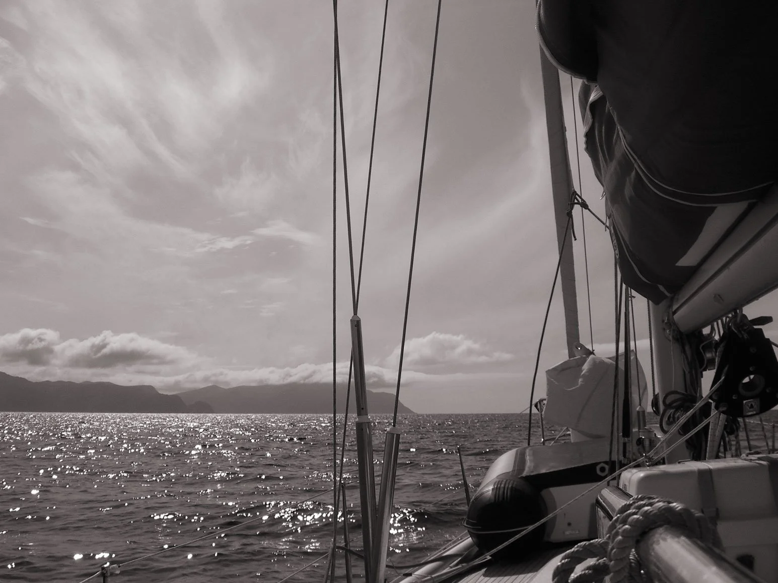 View from our sailboat's deck looking out over the ocean with a distant landmass and mountains, clouds, and sunlight reflecting on the water.