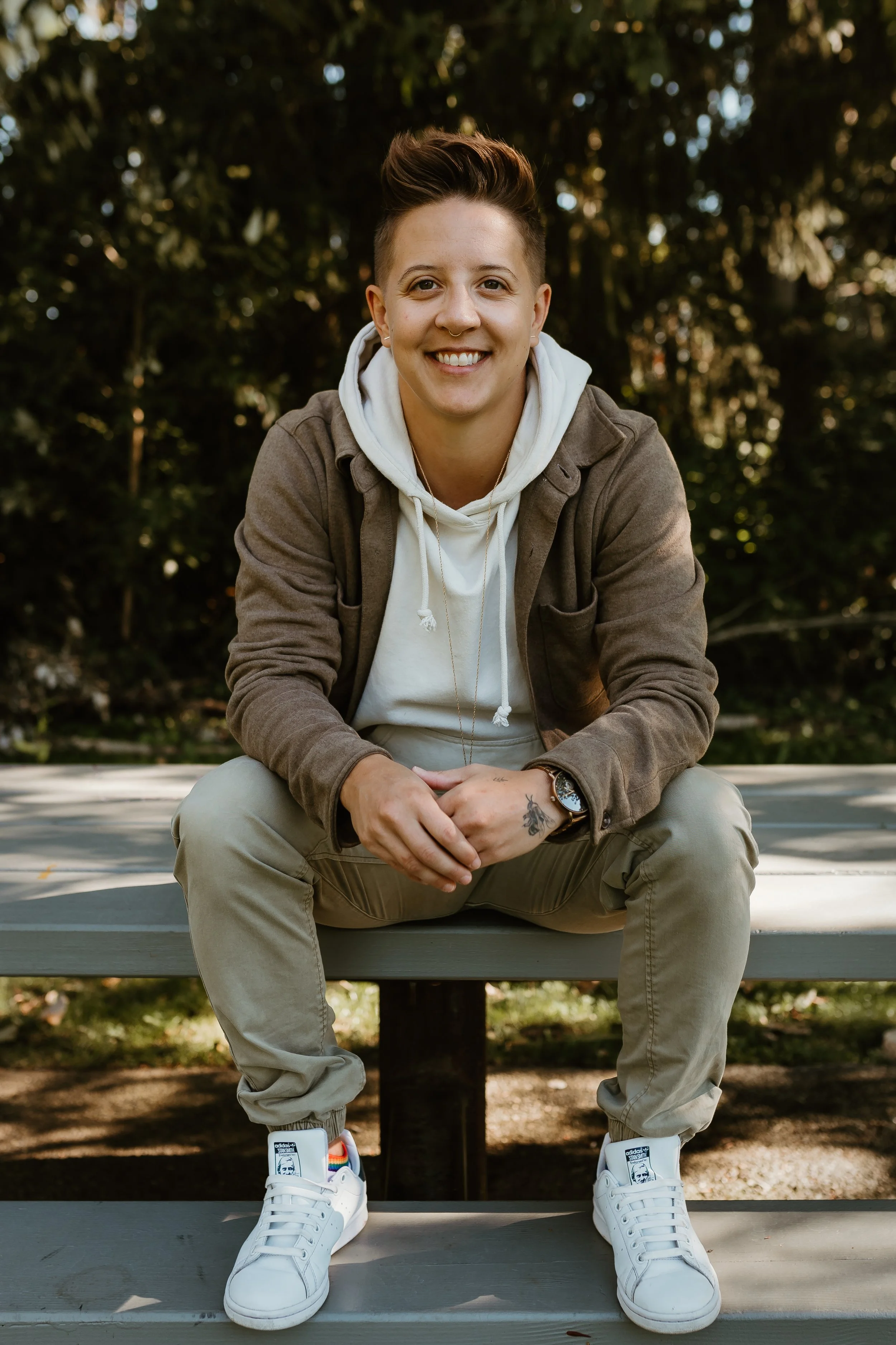 Erin Davis, LCSW & ADHD Therapist sitting on a bench outdoors in a park with trees in the background. They are wearing a light-colored hoodie, a brown jacket, beige pants, and white sneakers with a logo and rainbow-striped tongue.