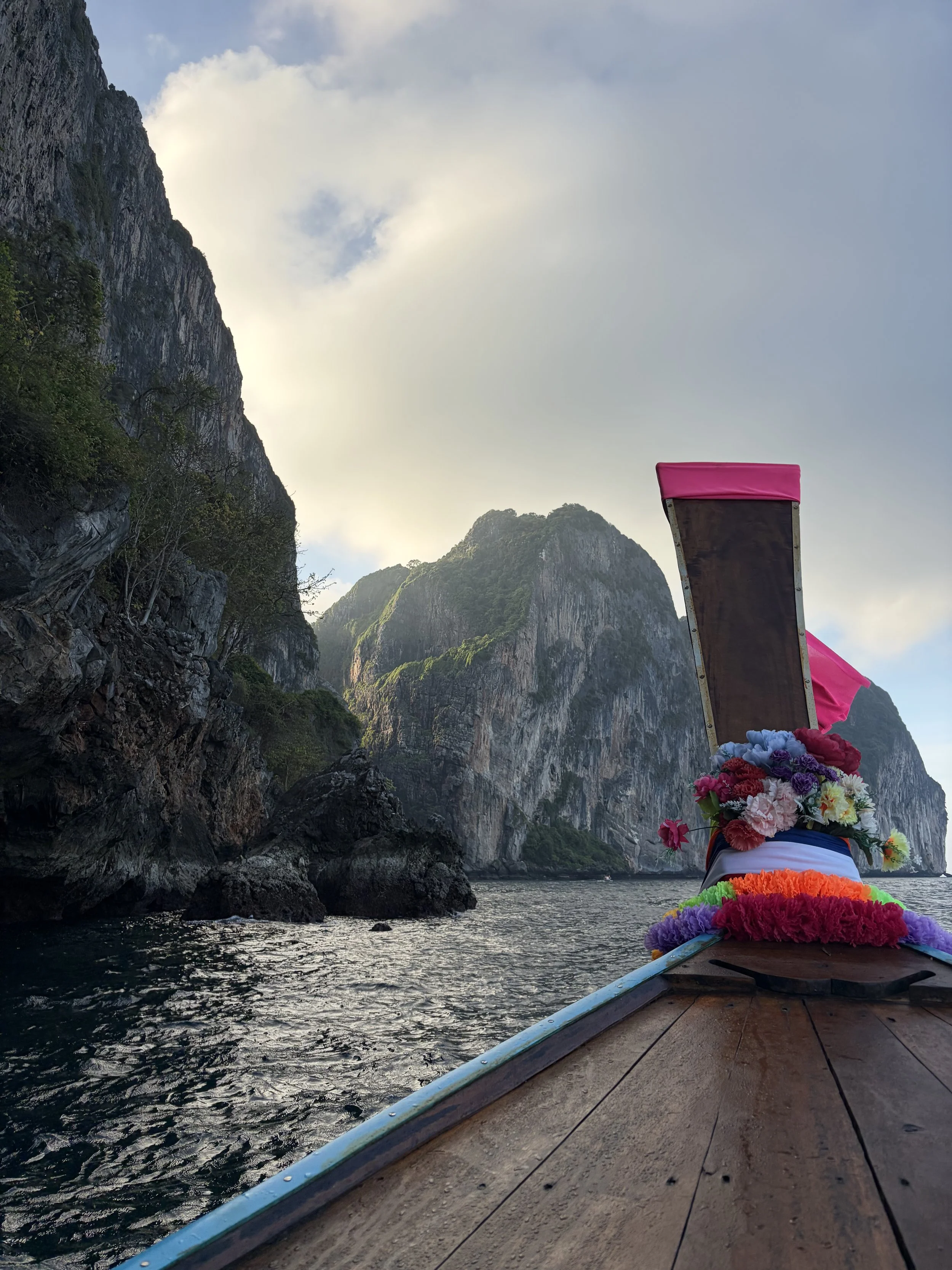 View from a boat with decorative flowers on a temple object in the water, surrounded by tall, lush mountains and a partly cloudy sky.