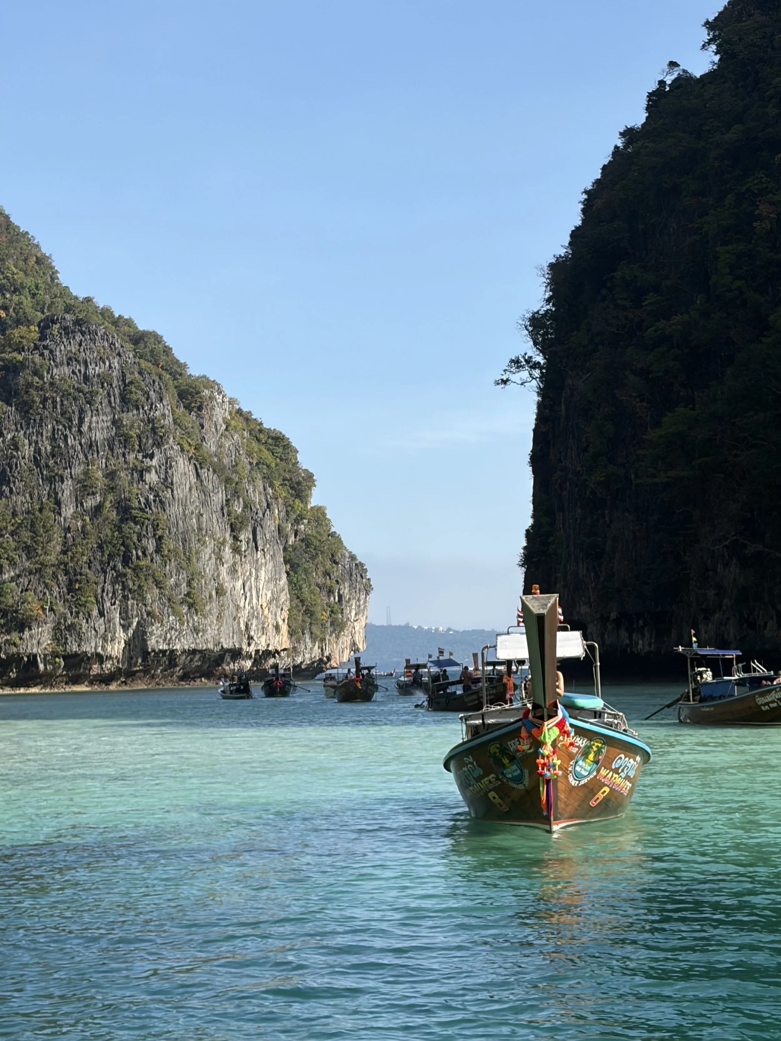 A speedboat with colorful decorations on the bow is floating on clear turquoise water in a narrow channel between two tall, green-covered cliffs. Several other boats are visible in the background, and the sky is clear.