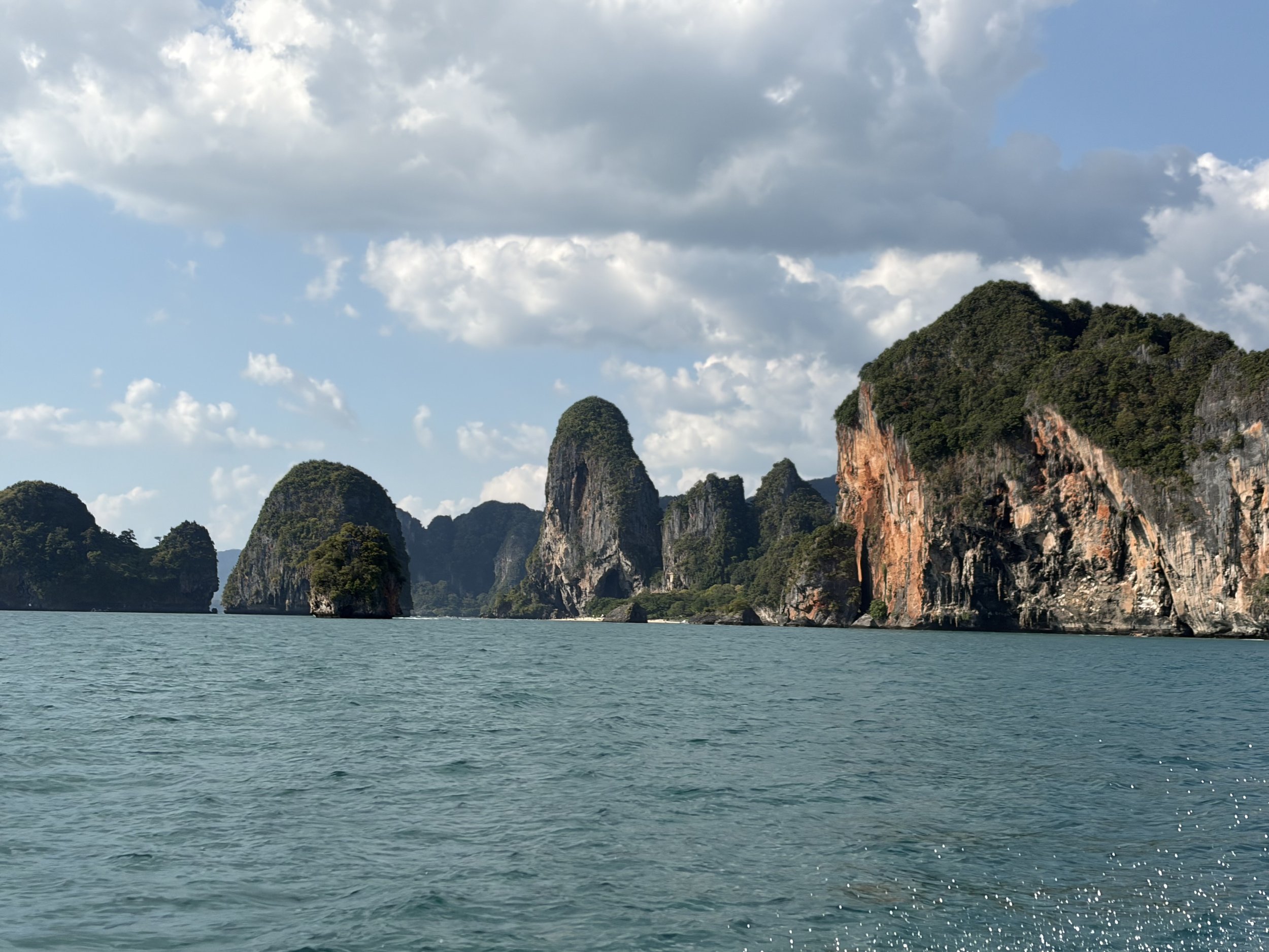 A scenic view of limestone karsts and cliffs rising from turquoise water under a partly cloudy sky.
