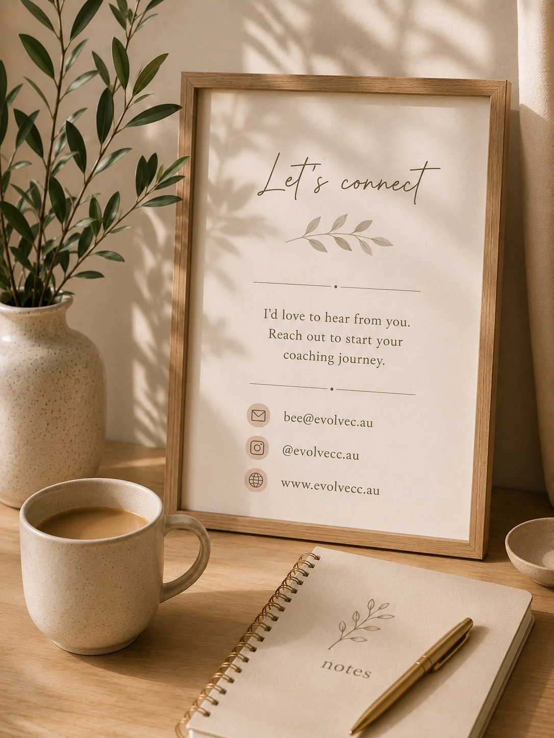 A framed sign on a wooden desk invites connections for coaching, displaying an email, Instagram handle, and website, alongside a potted plant, a coffee mug, a notepad with a leaf illustration, and a pen.