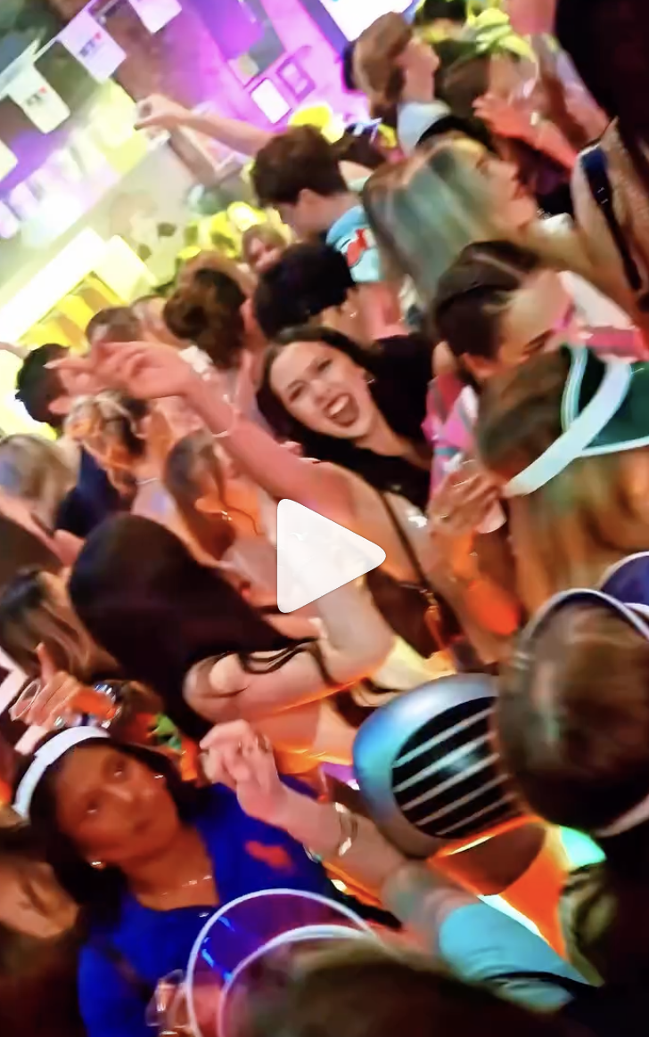 Large crowd of people at a lively party or celebration, dancing and with some wearing headphones, in a colorful, vibrant environment.