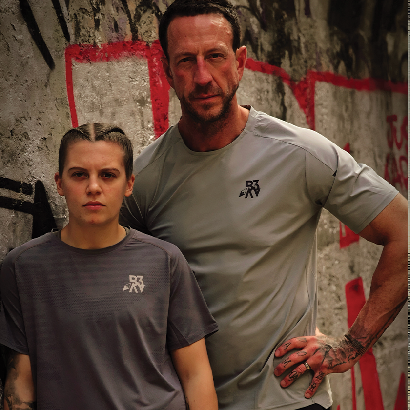 A young girl with braided hair and tattoos on her left arm, and a man with short dark hair, tattoos on his right hand, both wearing gray athletic shirts, standing against a graffiti-covered wall.