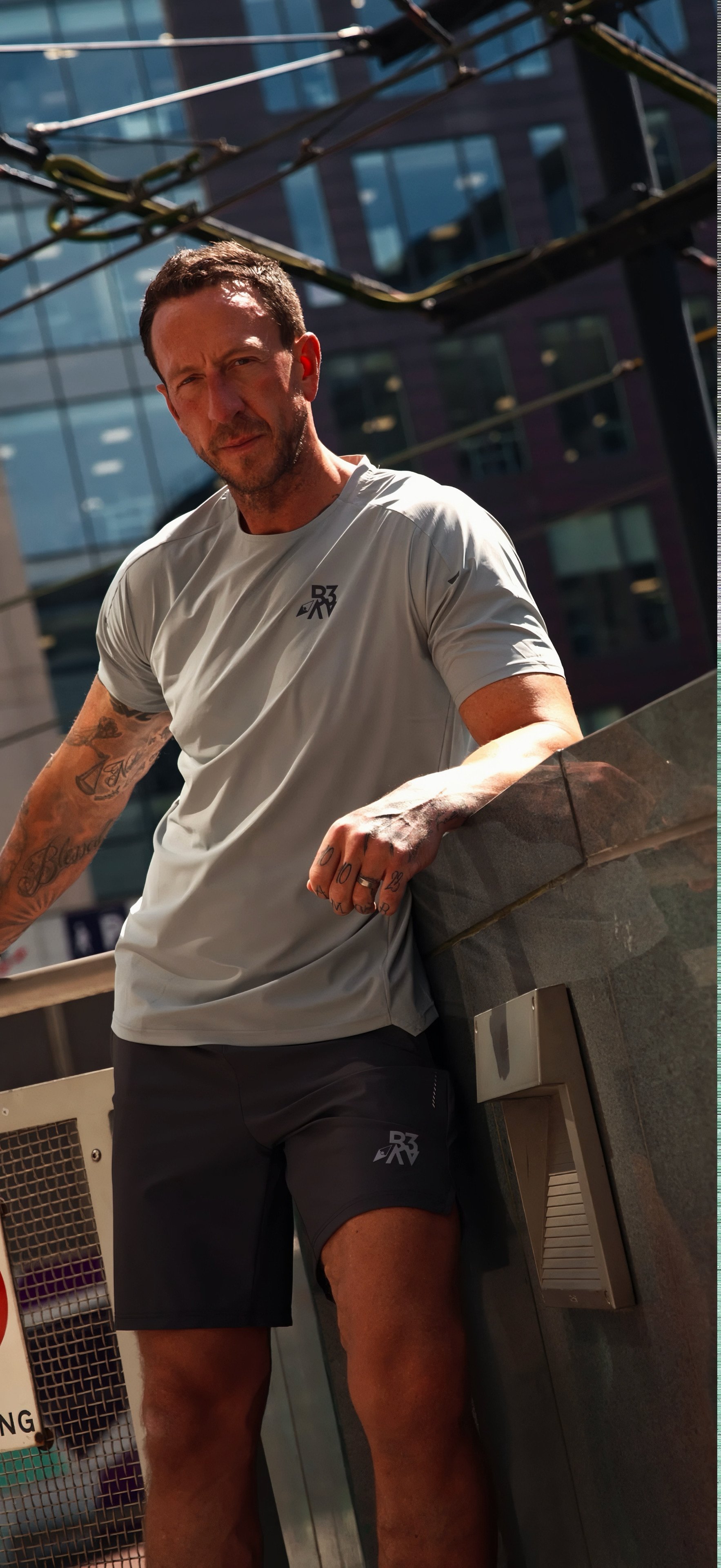 A man leaning on a railing outdoors with a modern glass building in the background, wearing a gray athletic shirt and black shorts.
