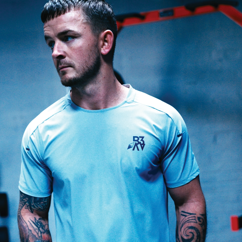 A man with short dark hair and tattoos on his arms wearing a light blue athletic shirt in a gym or training facility.