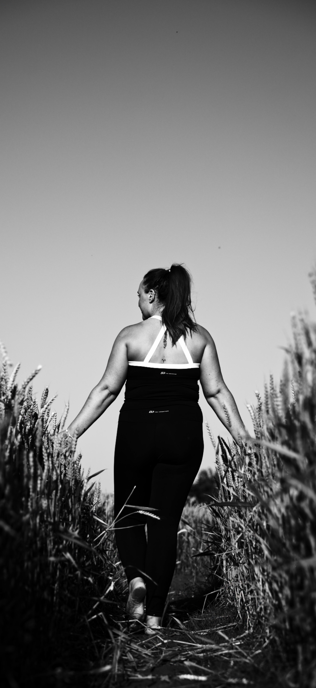 A woman in athletic clothing walking through a field of tall grass, with her back to the camera, in black and white.