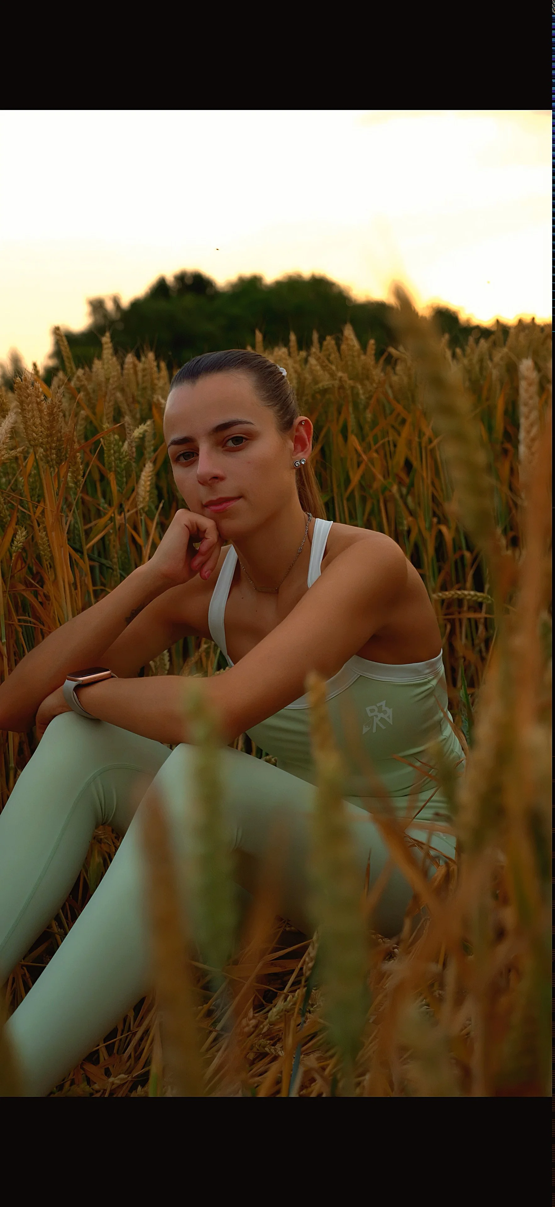 Young woman sitting in a field of wheat during sunset, wearing athletic clothing and jewelry.