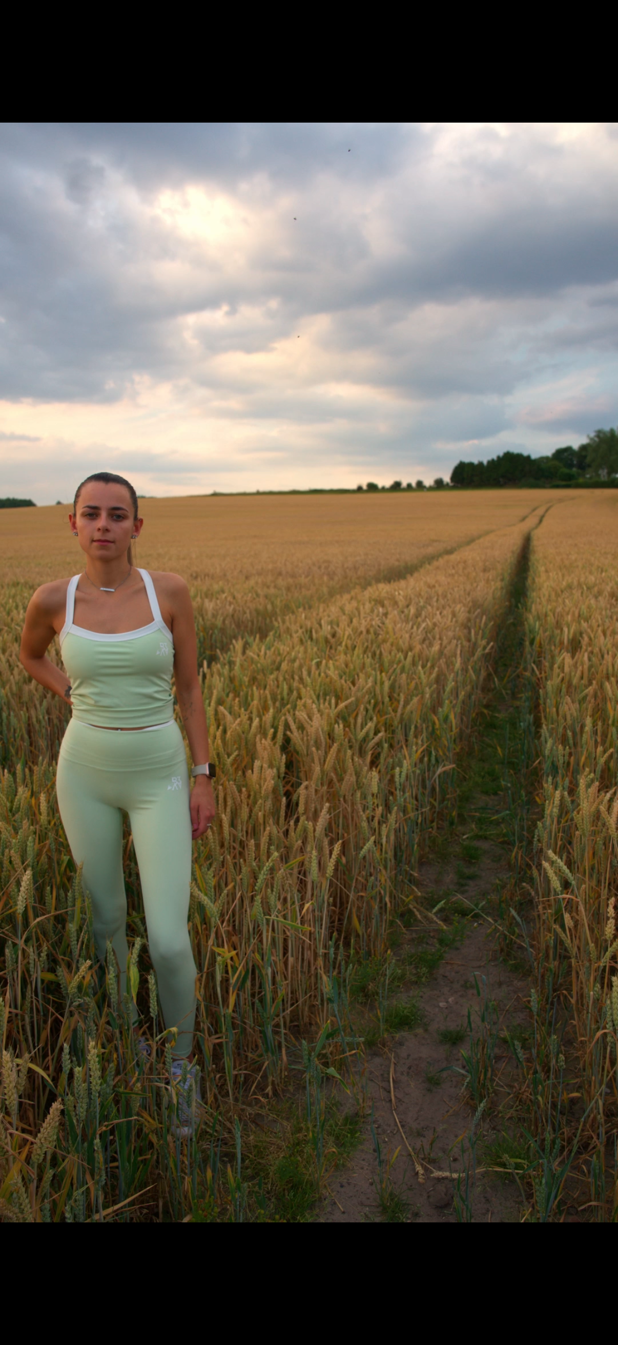 A young woman dressed in light yellow athletic wear standing in a wheat field during cloudy weather.