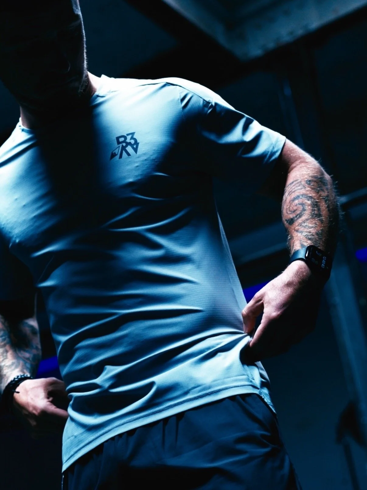 Close-up of a man wearing a white athletic shirt with a logo, flexing his left arm, showing a tattoo on his arm, in a dark indoor setting.