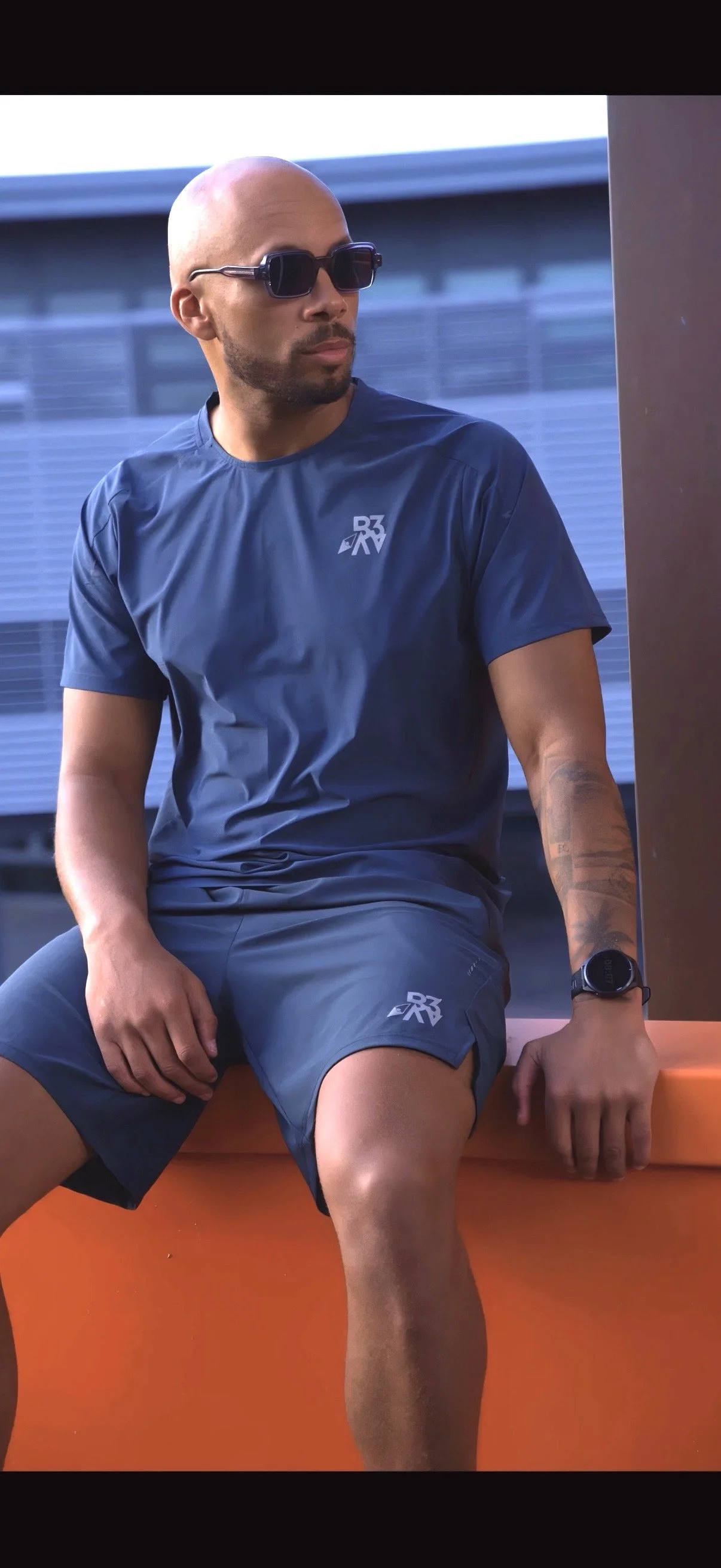 A man with a shaved head and beard wearing sunglasses, a navy athletic shirt and shorts, sitting on an orange surface, with a building in the background.