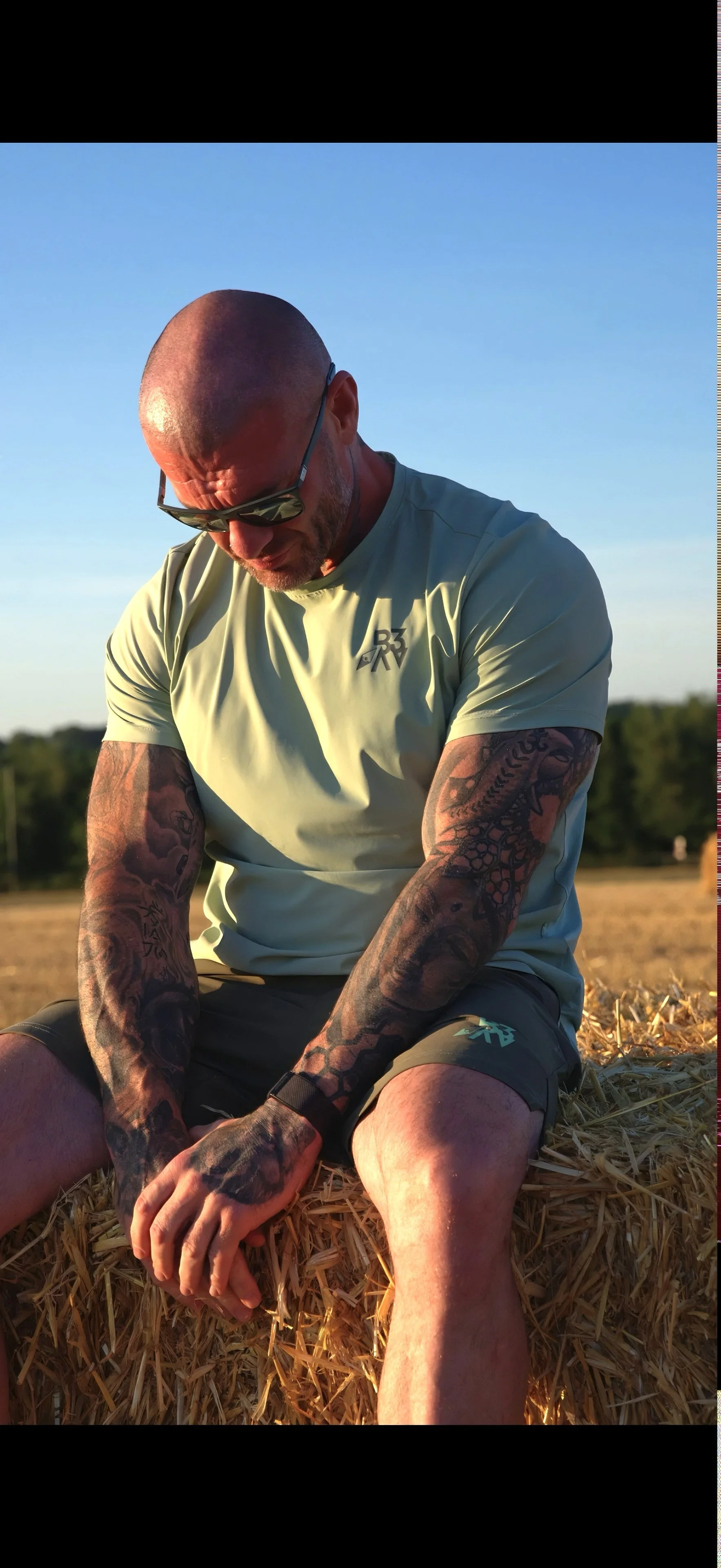 A muscular man with tattoos on his arms, wearing sunglasses and a yellow athletic shirt, sitting on straw in an outdoor field during sunset, with a clear blue sky and trees in the background.