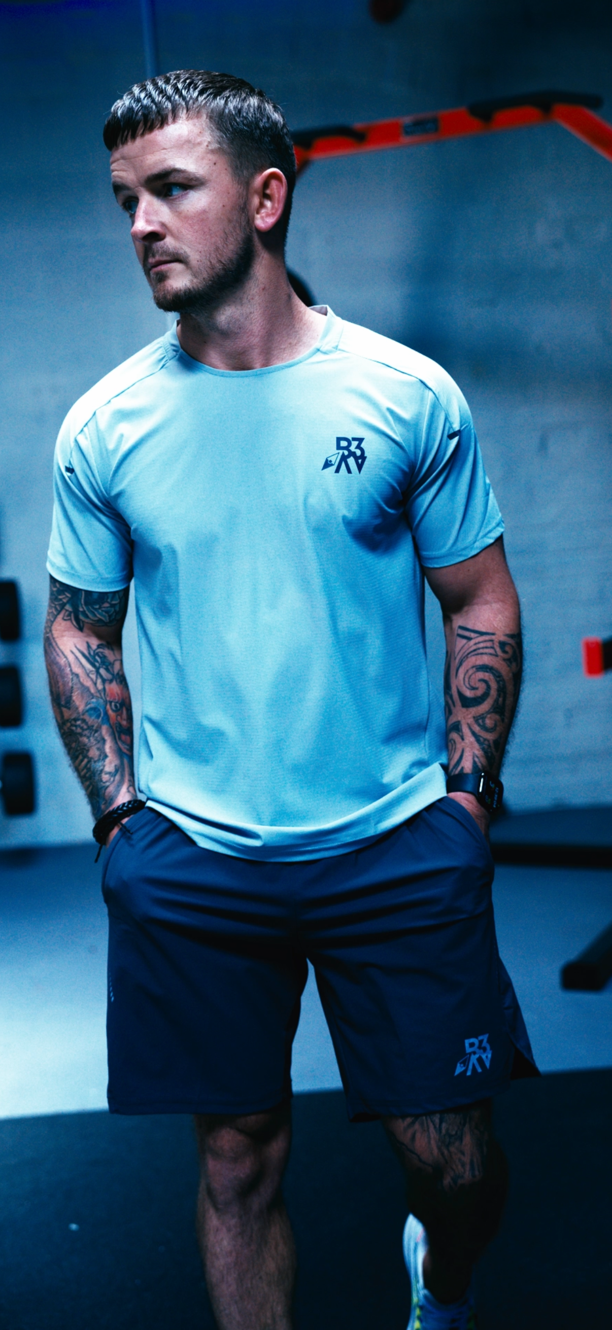 A man in athletic wear standing in a gym with his hands in his pockets, featuring tattoos on his arms and wearing a fitness tracker.