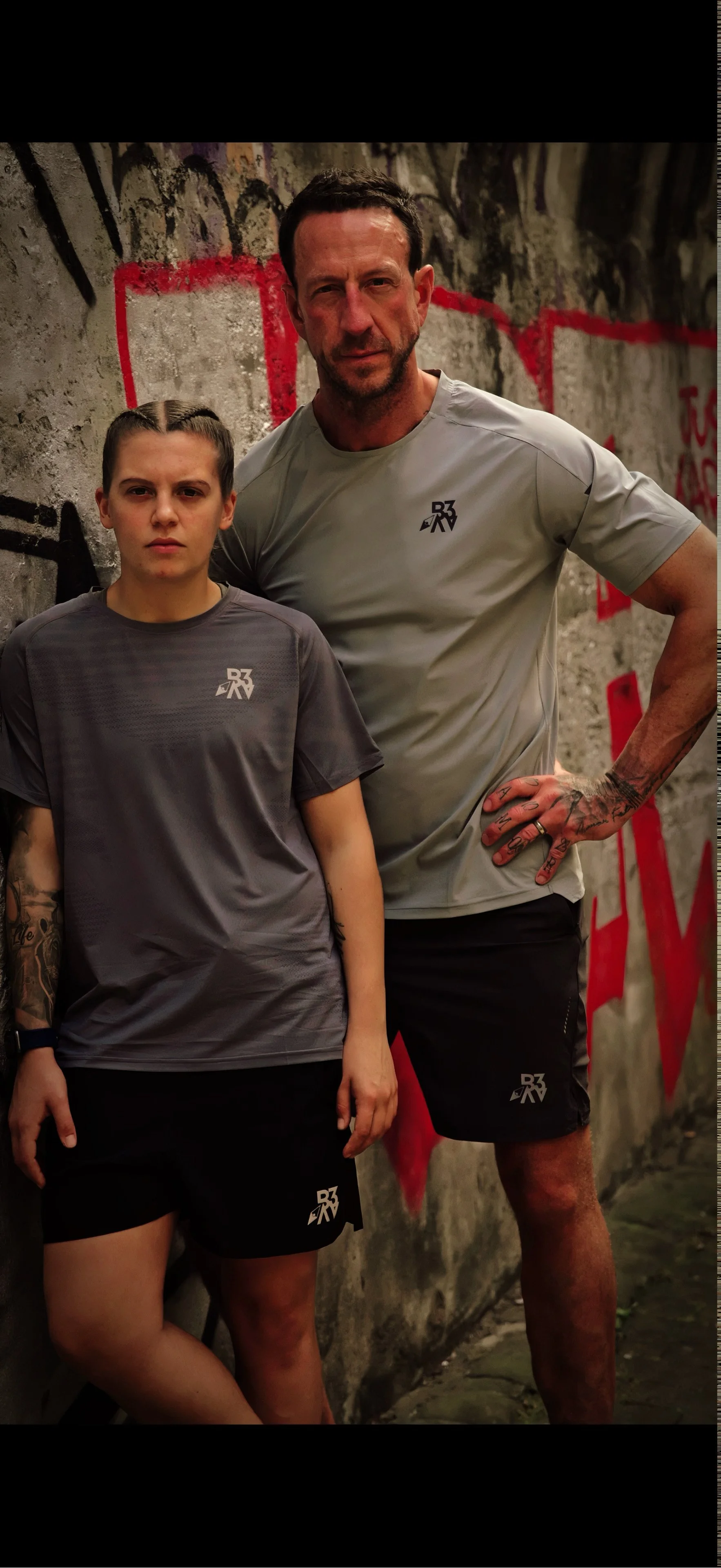 Two people, a young woman and a man, posing against a graffiti-covered concrete wall. Both are wearing matching athletic shirts and shorts with a logo, and the man has tattoos on his right arm and hand.