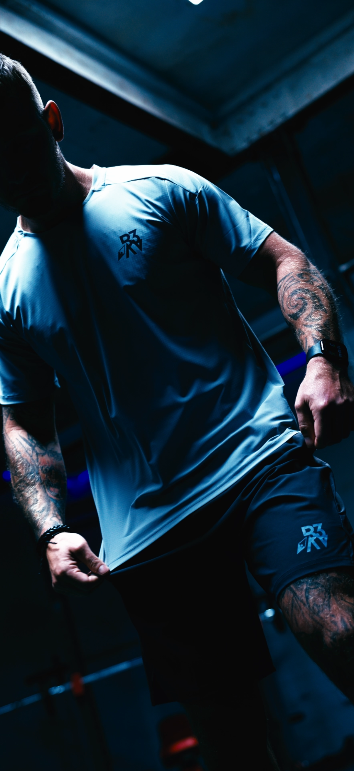 A man wearing athletic clothing, including a gray t-shirt with a logo and black shorts, is adjusting the bottom of his shirt in a dimly lit, gym-like environment with steel beams overhead.