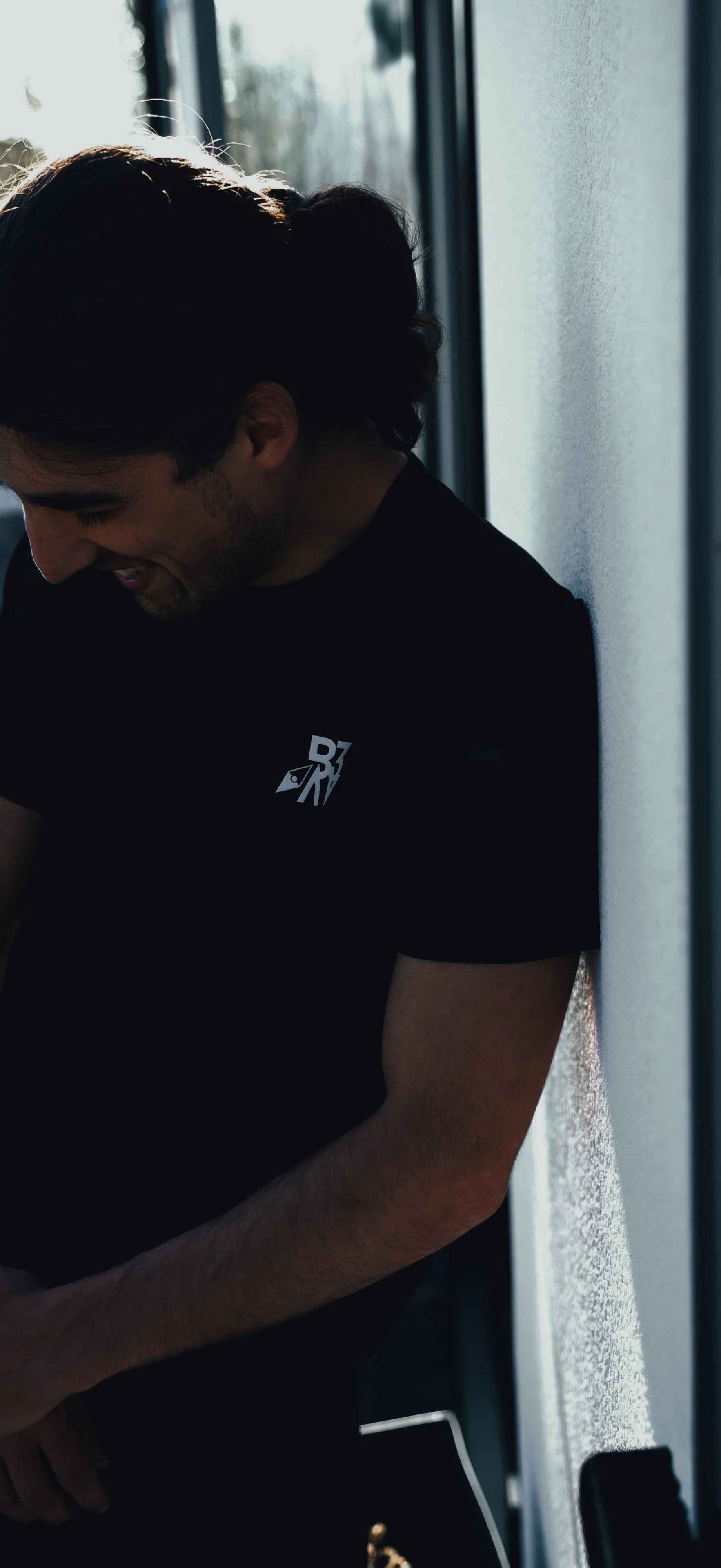 Young man laughing and smiling with head slightly bowed, wearing a black T-shirt with a small graphic on the chest, standing and leaning against a wall near a window with sunlight in the background.