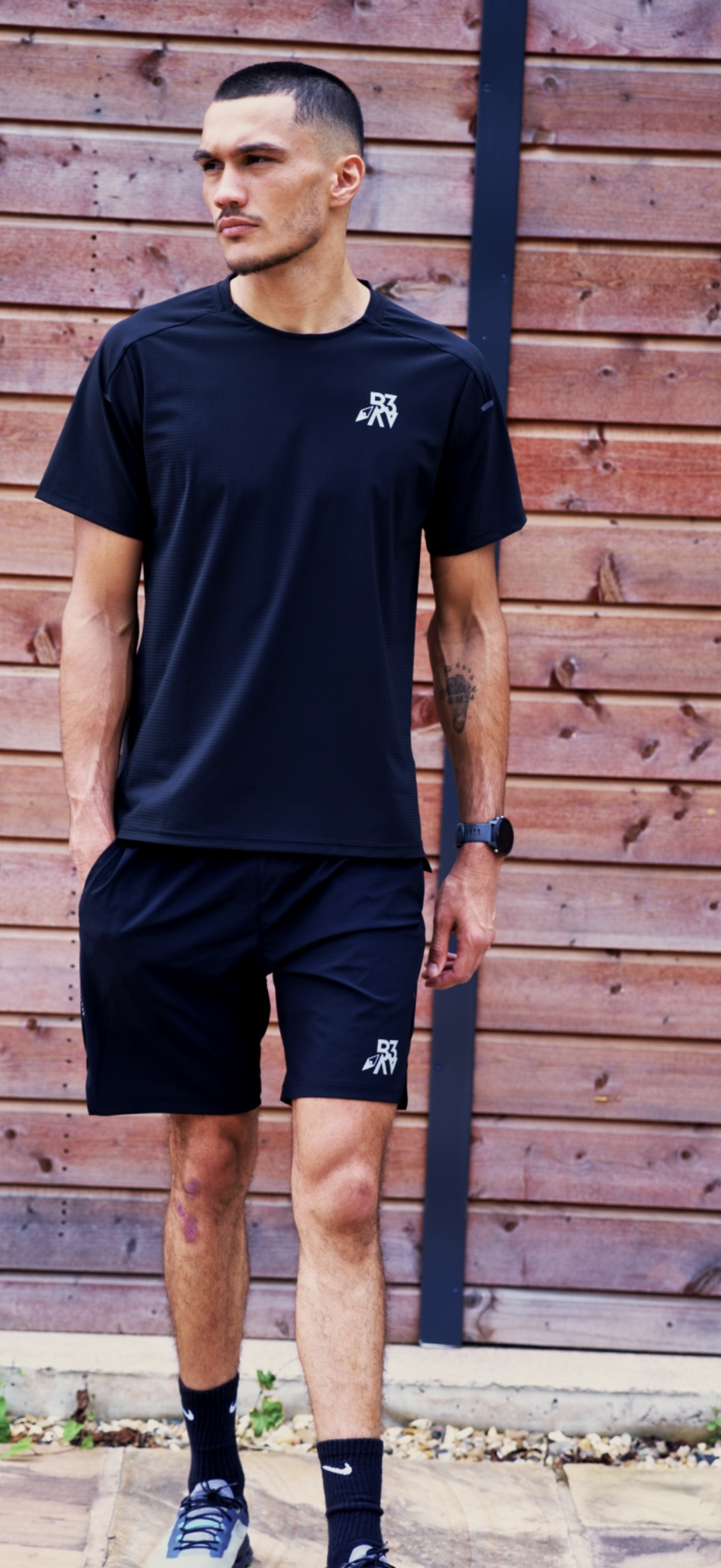A young man in black athletic clothing standing outdoors, leaning against a wooden fence.